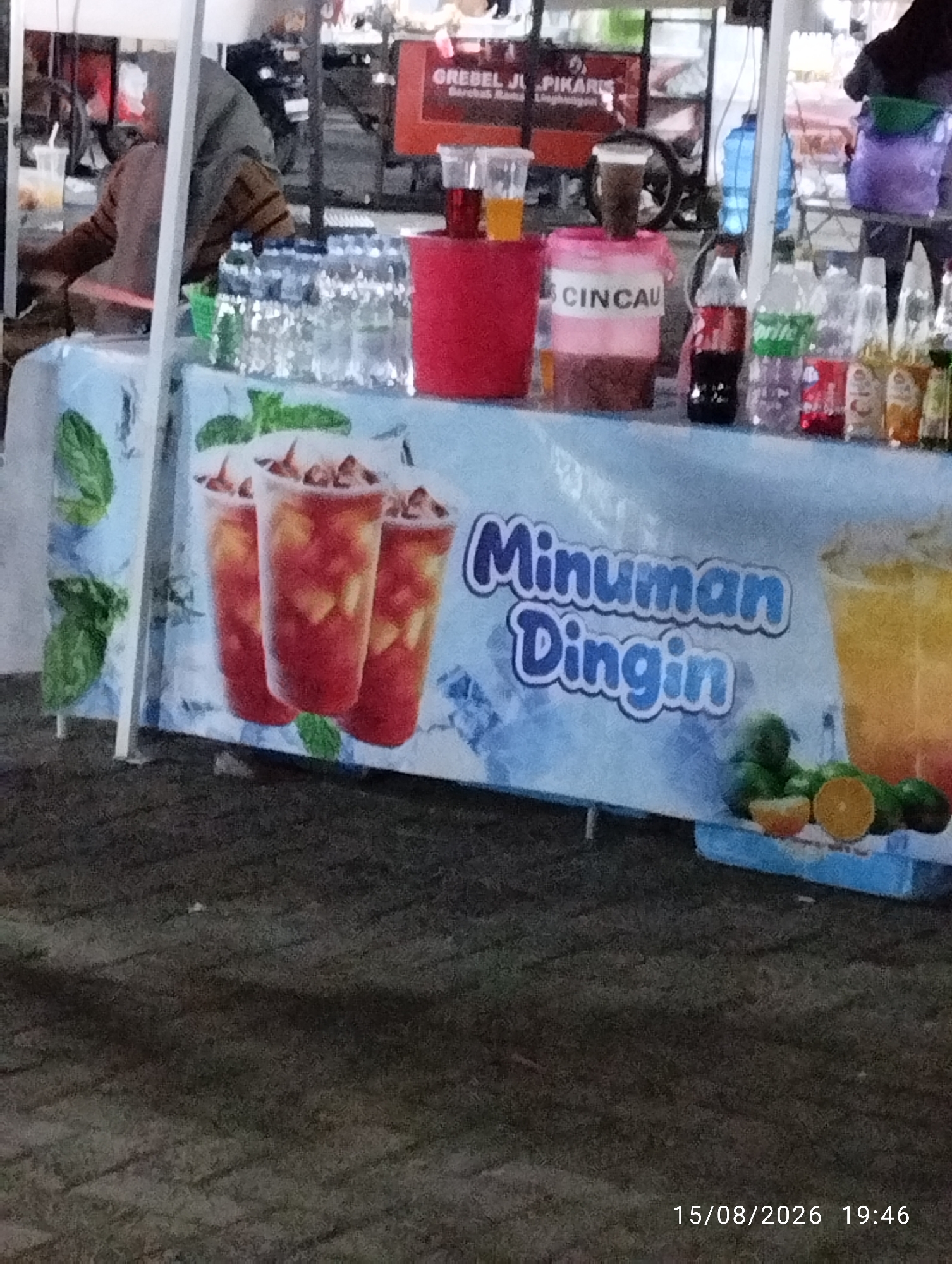 An outdoor vendor stall is depicted, displaying a variety of drinks and their ingredients. In the foreground, a light blue banner features a graphic of three iced beverages and the text "Minuman Dingin" which translates to "Cold Drinks". To the right of the banner, a tall glass of a layered orange and yellow beverage is shown, with sliced oranges and green fruits at its base. Behind this, several plastic bottles of water and soft drinks are arranged on a shelf, including a red Coca-Cola bottle and a green Sprite bottle. A pink cylindrical container with the word "CINCAU" printed on it sits to the left of these bottles, holding a dark substance. In the background, a person wearing a headscarf and a striped garment is partially visible, appearing to be attending the stall. The stall itself has a white frame and a blue tarp. The ground in front of the stall is paved with dark, textured material, and a timestamp reading "15/08/2026 19:46" is visible in the lower right corner.