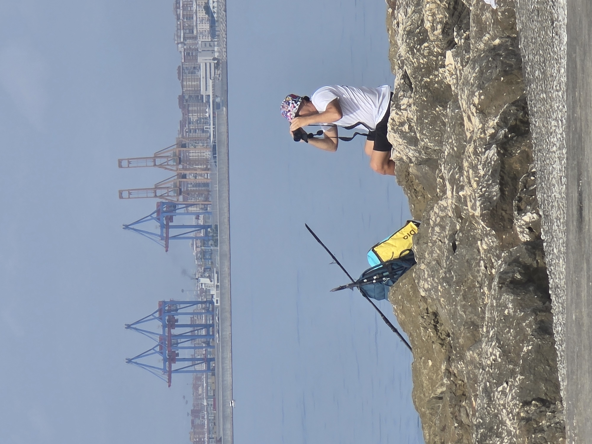 A man wearing a white t-shirt, black shorts, and a colorful bucket hat is sitting on a rocky shoreline, using binoculars to look out over the water. Behind him, a brightly colored yellow and blue bag is propped up against some sticks on the rocks. In the background, a large port with numerous cranes is visible across the water, with a city skyline beyond. This scene is likely located at the port of Málaga, Spain. The water is calm and blue, and the sky is overcast.