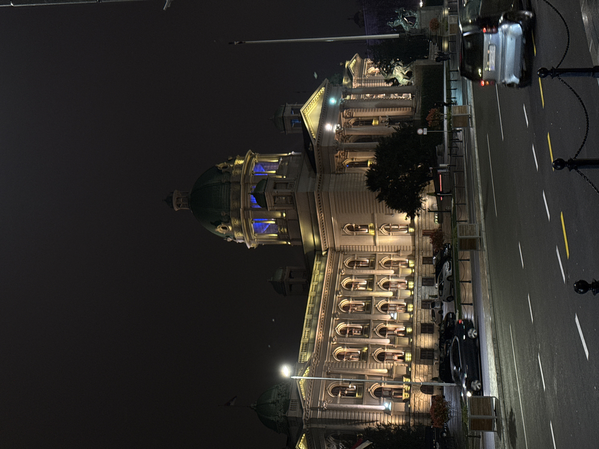 At night, the National Assembly of Serbia in Belgrade is illuminated with golden lights, emphasizing its architectural details. The building features a dome, visible windows, and classical columns. Along the adjacent street, cars are parked alongside a sidewalk lined with trees. The road is marked with white and yellow lines, and decorative bollards line the edge.