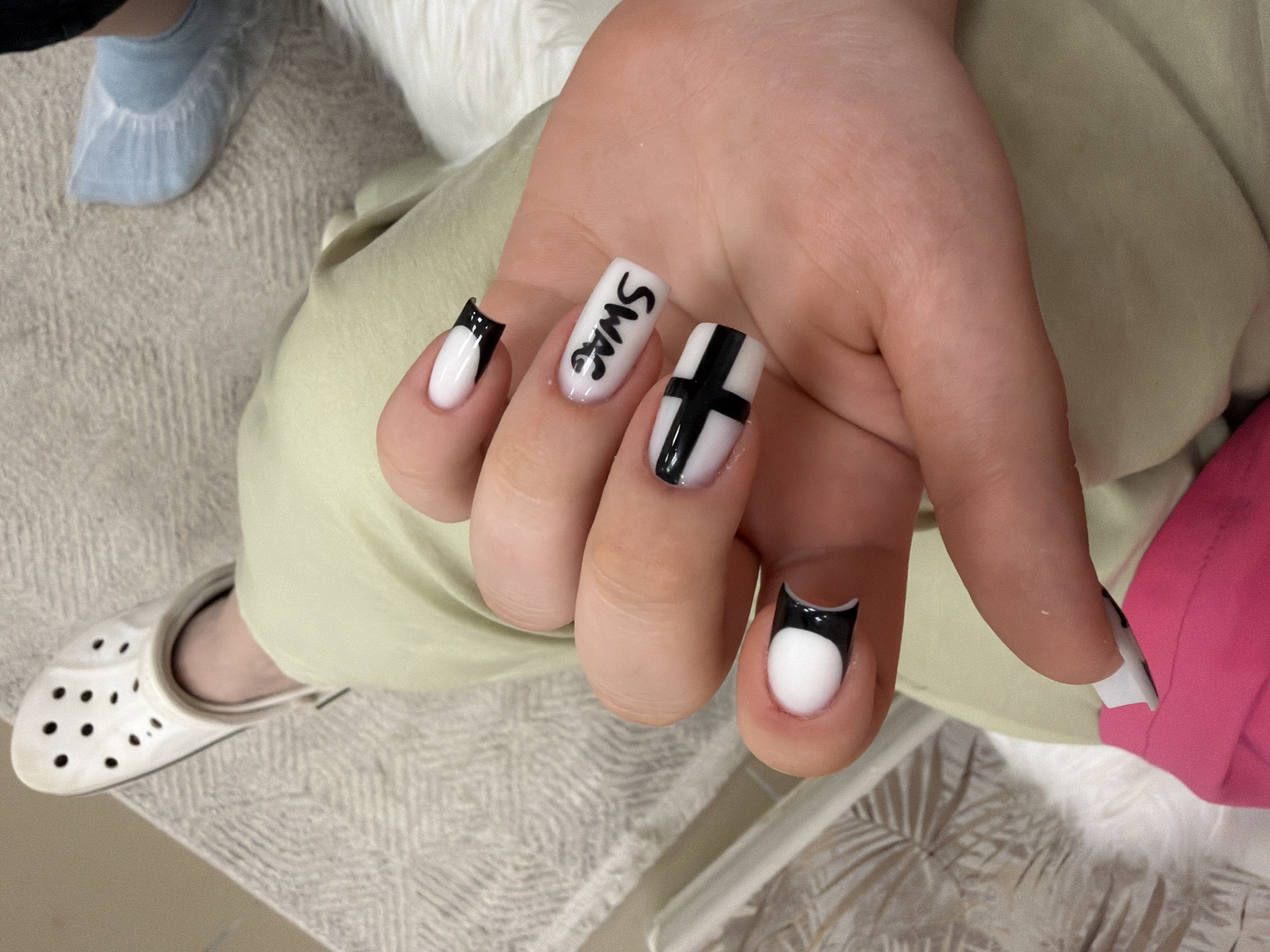 The image shows a close-up view of a person's hand with white and black nail art. The nails are short and squared. The thumb has a black cross design. The index finger has the word "swag" written in black letters. The middle finger also has a black cross design, and the ring finger has a black french tip. The person is wearing light green pants. In the background, a white Croc shoe is visible on the left, and a light blue sock on the right. The environment appears to be indoors, with a textured rug beneath the feet. There is no indication of the location being in the city of Perm, Russia.