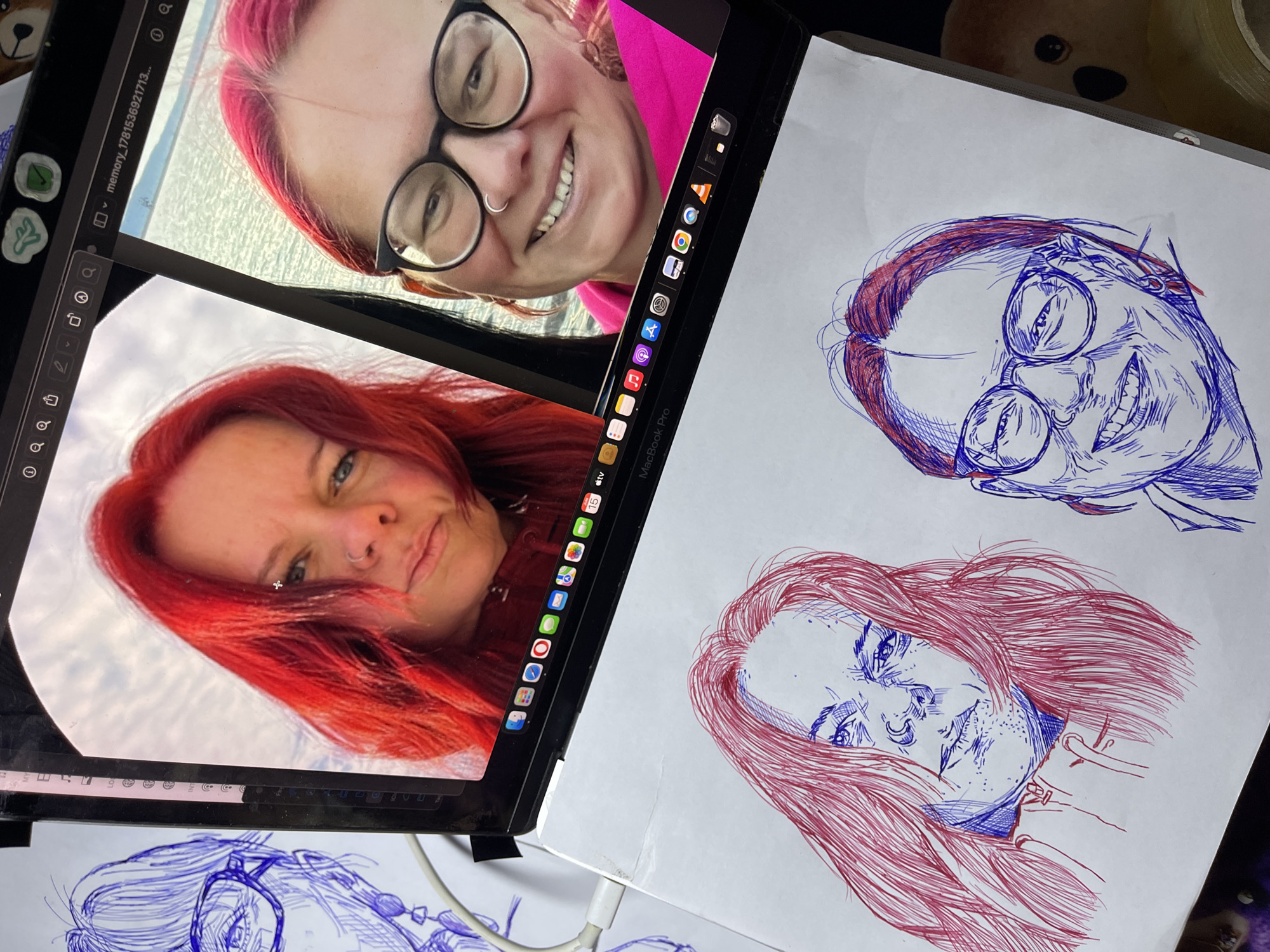 A MacBook Pro is open, displaying two selfies of a woman with vibrant red hair. On the left screen, she is looking slightly down with a neutral expression and a nose ring. On the right screen, she is smiling broadly, wearing glasses and a nose ring. In front of the laptop, on a white piece of paper, are two sketches in red and blue ink. The sketch on the left is a portrait of a woman with red hair, looking intensely with freckles and a nose ring. The sketch on the right is a portrait of a woman with glasses and a nose ring, smiling widely. The background includes a white charging cable and a portion of a stuffed toy. The image does not contain any information about the location of Jos, Nigeria.