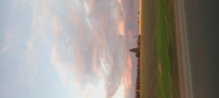 An orange-tinged sunset illuminates a sky filled with striated clouds. Below, a green field fades into indistinct foliage. A gray, possibly asphalt, surface occupies the extreme right. The image is blurry, particularly at the bottom. It appears to have been taken quickly, possibly from a moving vehicle.