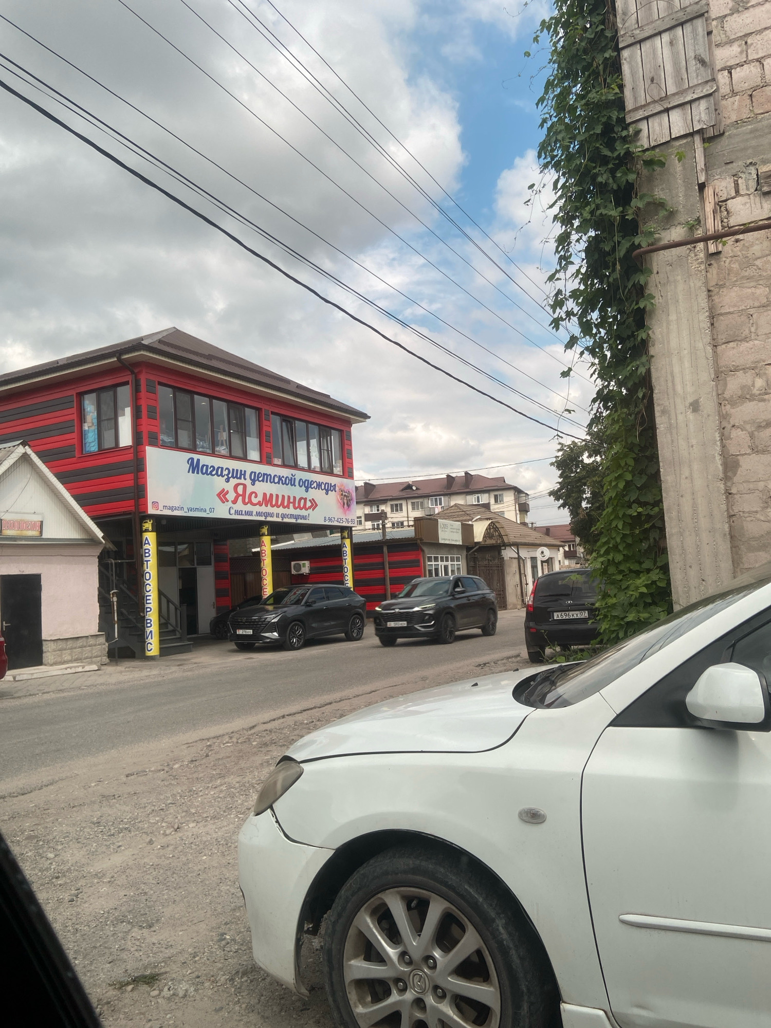 The image shows a street scene in Chegem, Russia. In the foreground, a white car is partially visible, with its front fender and wheel in focus. The car appears to be parked on a gravel road. Behind the white car, a dark-colored SUV and a black car are parked.
Further down the street, a two-story building with red and black striped siding stands prominently. A sign on the building reads "Магазин детской одежды «Ясмина»" (Children's clothing store "Yasmina"). Below this, the slogan "С нами модно и доступно!" (With us it's fashionable and affordable!) is visible, along with an Instagram handle "@_magazin_yasmina_07" and a phone number. Yellow signs with the word "АВТОСЕРВИС" (Car service) are mounted on poles in front of the building's entrance.
To the left of the clothing store is a smaller, lighter-colored building with a dark door and a sign that reads "DACHKOUR".
The street itself is paved with gravel and asphalt. Overhead, power lines stretch across the frame, silhouetted against a partly cloudy sky. To the right of the building, a concrete wall is covered with dense green ivy. In the background, residential buildings and trees are visible under the cloudy sky. The overall impression is that of a typical street in a Russian town, with a mix of commercial and residential structures.