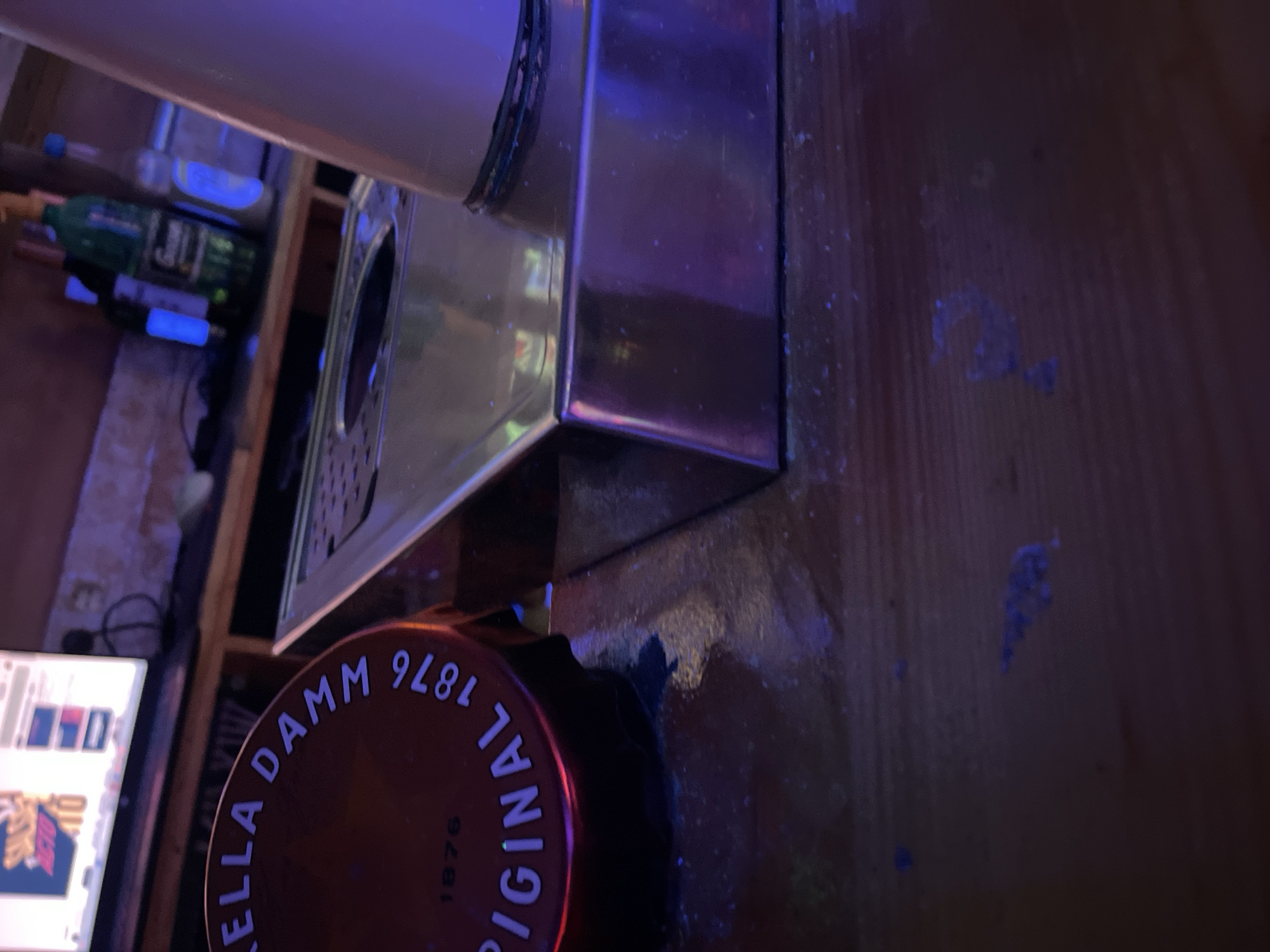 The image depicts a close-up, low-angle view of a wooden bar surface in what appears to be a dimly lit establishment, possibly a bar or pub. In the foreground, a metallic object with a rectangular base and a curved top is visible, with a hole in its center. To the left, a round, reddish-brown object with white lettering that reads "STELLA DAMM 1876 ORIGINAL" is positioned, suggesting it's a cap or lid of a beverage container. The background shows a shelf with several bottles, including one green bottle with a yellow cap, and a monitor displaying what seems to be a digital screen with some text and graphics. The overall lighting is dim, with a purple hue suggesting ambient colored lights, common in nightlife venues. The surface of the bar appears slightly wet or sticky in places. The context of Barcelona, Spain is not visually evident in the image itself.