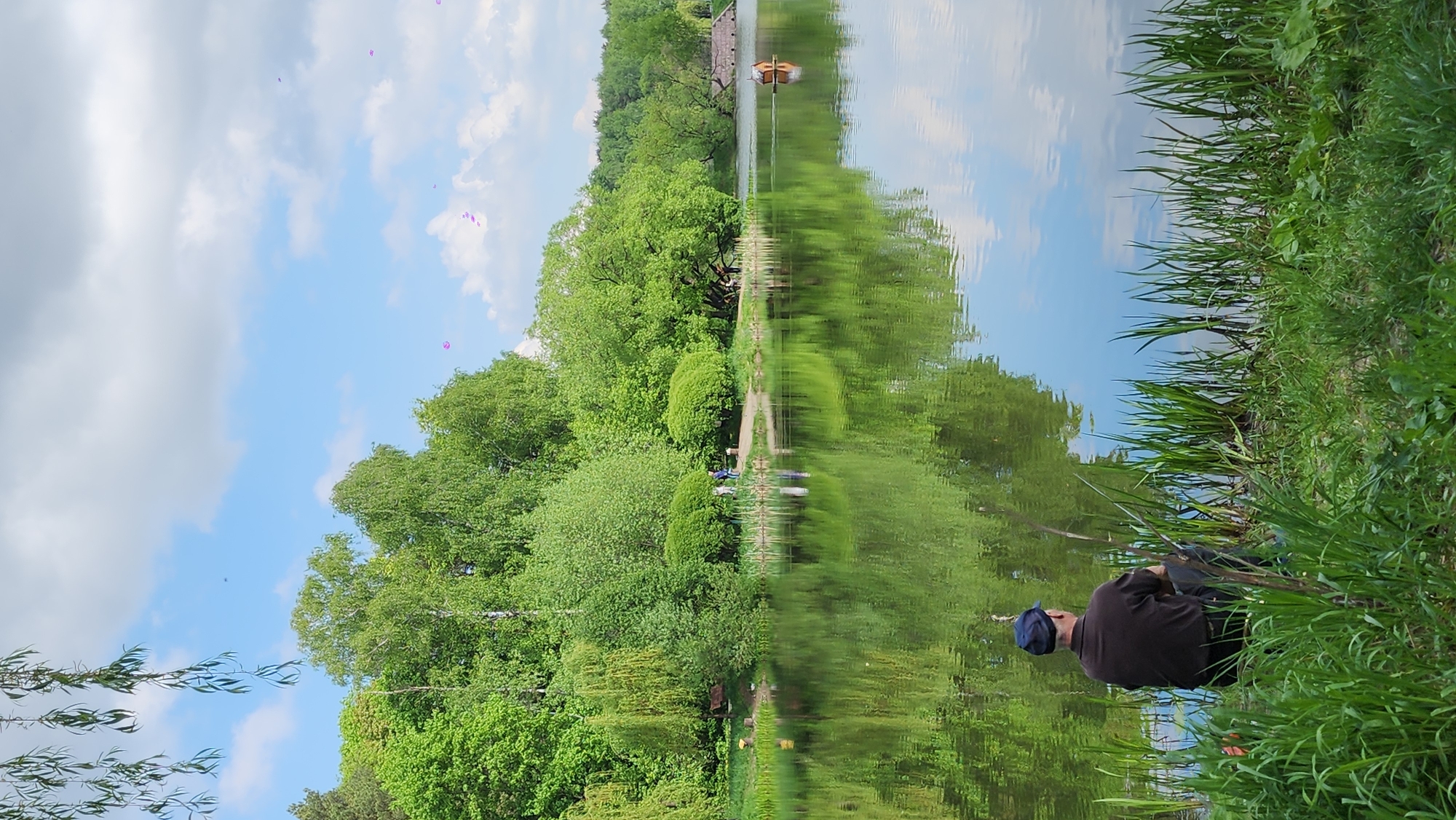 A landscape shot of a lake or large pond in Khimki, Russia. The water reflects trees, sky, and structures. A man in dark clothing and a blue cap sits on the grassy bank, possibly fishing. Tall grass obscures part of the lower right. Purple balloons float in the upper left. A small boathouse or dock appears on the other side of the water with a small boat. The sky is blue with white clouds.