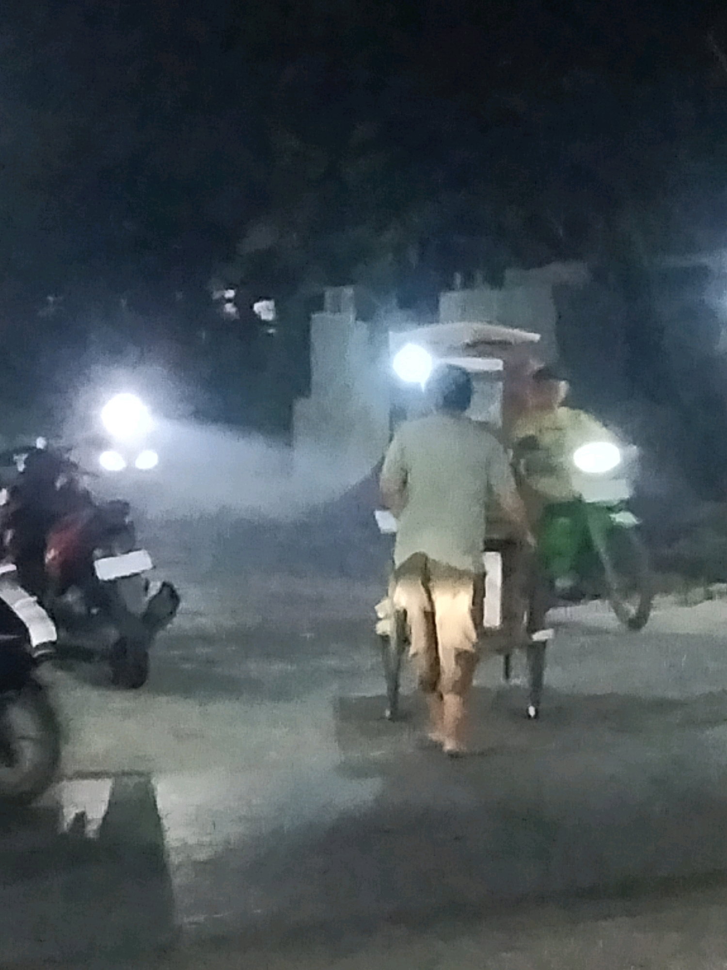 This is a night scene photograph taken in Pisangan Jaya, Indonesia, likely on a street. Two individuals are depicted. The foreground features a person from behind, wearing a light-colored short-sleeved shirt and light-colored shorts or pants, pushing a cart. The cart appears to have a lighter-colored structure on top and is supported by dark-colored legs with wheels visible. To the right and slightly behind, a second person is seated on a motorcycle, which is also dark-colored and has a bright headlight. This individual wears a light-colored shirt and green pants. The motorcycle is in motion, indicated by the blur of its wheels and a bright light from its front. The background is dark and indistinct, with some faint illumination suggesting buildings or structures. Several motorcycles are parked or moving in the background, with some exhibiting bright headlights and taillights, and one emitting a visible puff of smoke or steam. The ground surface appears to be asphalt or pavement.