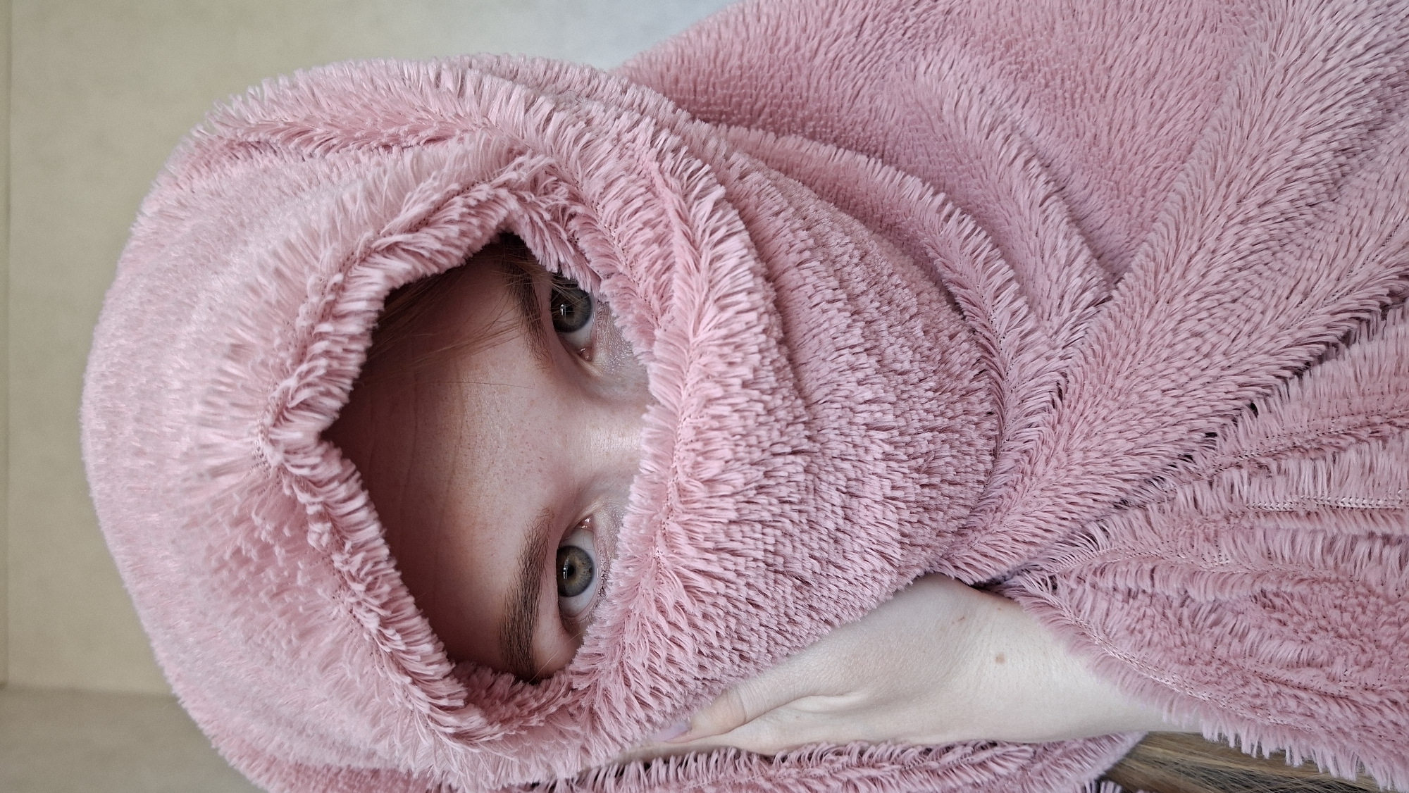A person is mostly enveloped in a soft, pink, furry fabric, possibly a blanket or a hooded garment. Only the face, from the forehead down to the upper lip, is visible between the folds of the fabric. The eyes are open and looking directly at the camera. A hand is visible at the bottom of the frame. The background is a plain off-white or light neutral color. The location is irrelevant since it cannot be determined from the image.