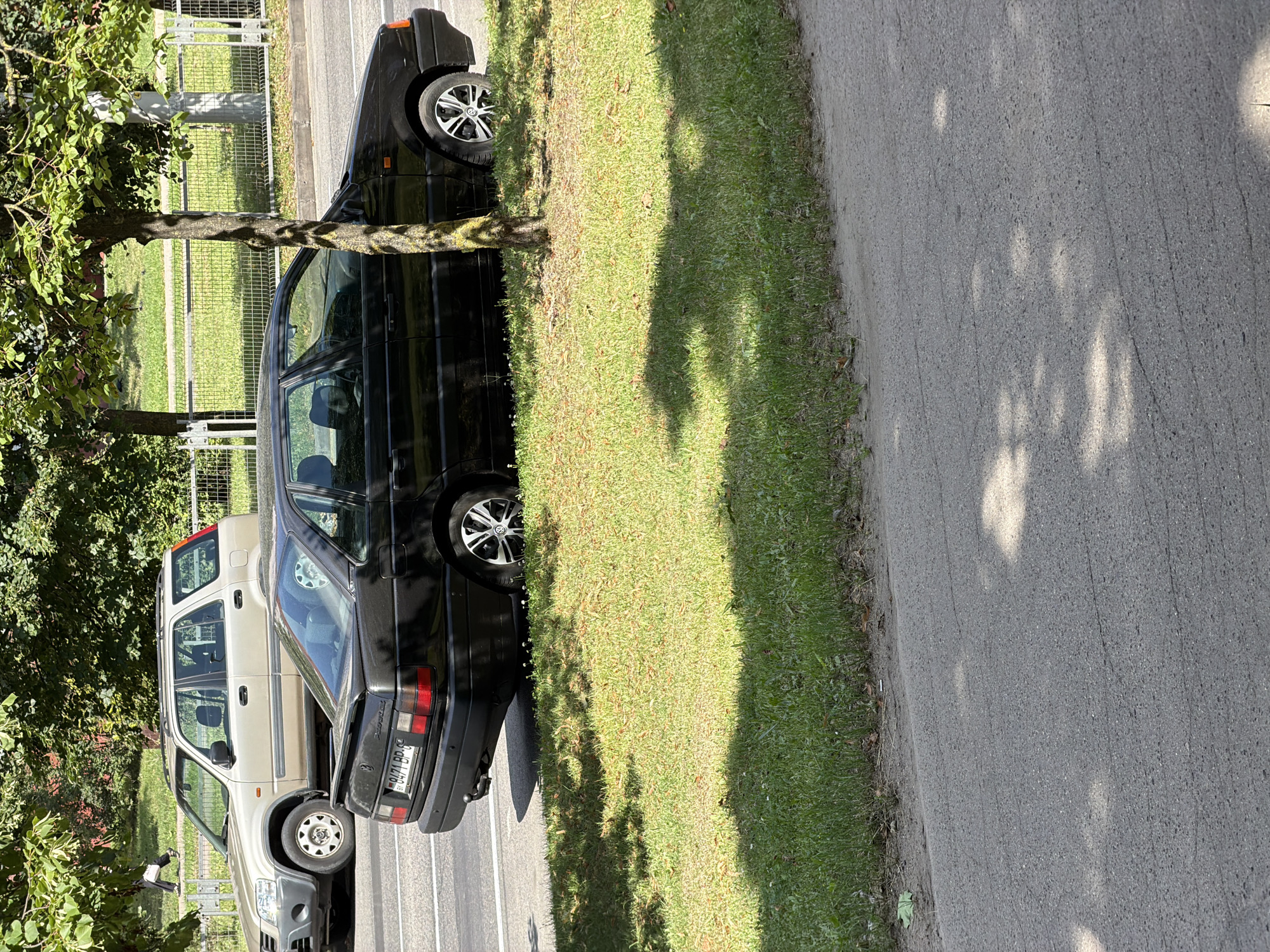 An outdoor, eye-level shot captures a black sedan parked on the right side of a street, partially obscured by a tree in the foreground. The car is positioned next to a patch of grass and a sidewalk, with a light-colored SUV parked further down the road in the background. Behind the vehicles, a metal fence runs along the edge of a grassy area, and trees with green foliage are visible, suggesting a sunny day. The license plate of the black sedan reads "0471 BP-6". The scene appears to be in a residential or urban area.