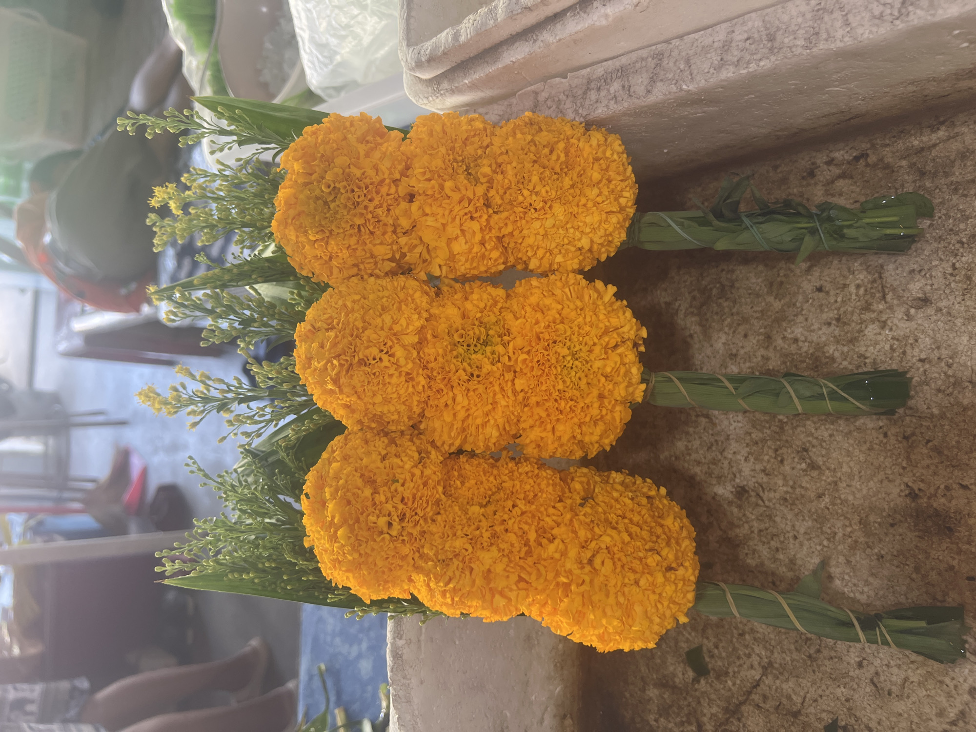 In an outdoor market setting in Ko Samui District, Thailand, three elaborately arranged bouquets of bright orange marigolds are presented. Each bouquet consists of multiple marigold flowers clustered together and bound with green foliage, tied with thin, light-colored string. The stems are wrapped in green leaves, secured with the same type of string. The marigolds are accompanied by delicate green ferns and small yellow flowers, adding a complementary textural and color contrast. The bouquets are arranged in a row on a weathered, speckled surface, possibly a countertop or stall. In the background, out of focus, are glimpses of people and market activity, including legs of individuals sitting or standing, and hints of market stalls and structures. The overall scene suggests a vibrant, possibly spiritual or celebratory, marketplace.
