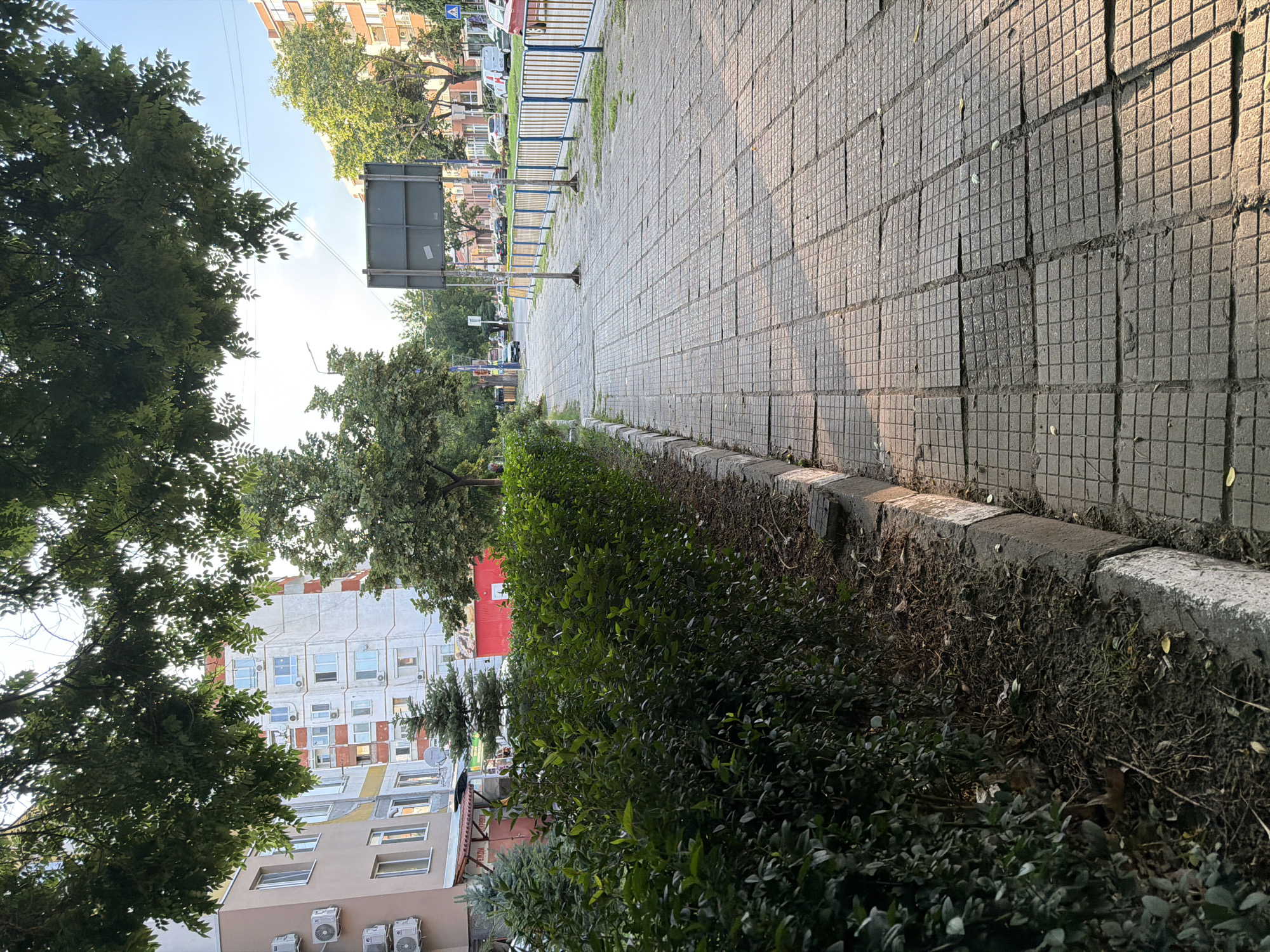 The image depicts a sidewalk in Pazardzhik, Bulgaria, characterized by worn square concrete tiles. A well-maintained green hedge runs along the left side of the sidewalk, separating it from a building with air conditioning units visible on its facade. Beyond the hedge, more foliage and trees are present. On the right side of the sidewalk, a yellow and blue metal fence encloses a grassy area, beyond which is a street with parked cars and other buildings. The sky is clear and blue, with sunlight casting shadows on the sidewalk, suggesting it is daytime.