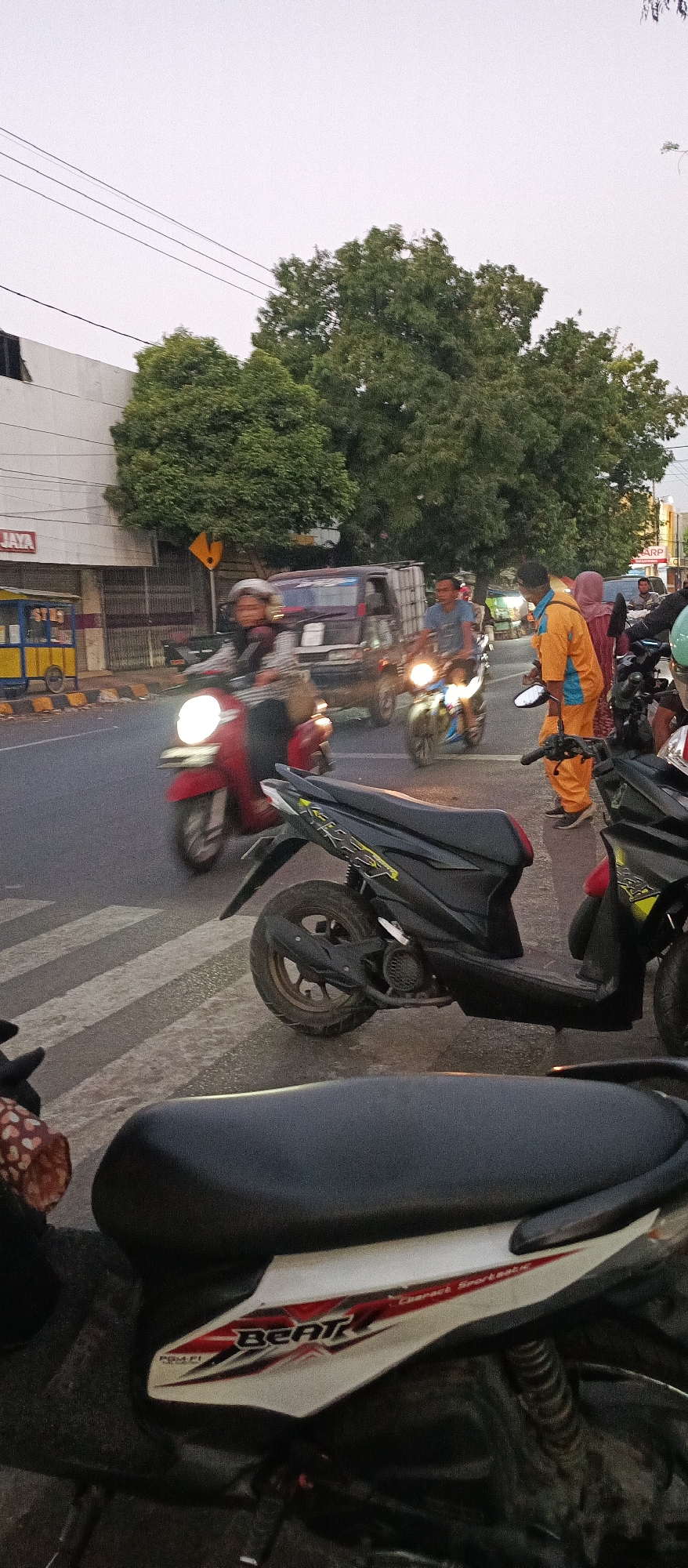 This vertical, medium-angle, outdoor photograph captures a street scene in Ardirejo, Indonesia, likely during the late afternoon or early evening due to the subdued lighting. The foreground is dominated by the rear portions of several parked scooters. A black scooter is prominently featured, with a white and red "Beat" graphic on its side panel. To its left, a portion of another scooter with a patterned seat cover is visible.
The midground depicts a busy street intersection with a pedestrian crossing. Several motorcycles are in motion. A red scooter with a rider is approaching the crossing from the left, its headlight illuminated. Behind it, a black van with a cargo box is moving forward. Another motorcycle, ridden by a person in a blue shirt, is also moving through the intersection, its headlight on. To the right, a person in an orange uniform, possibly a street cleaner or worker, stands near the roadside, interacting with another individual. Further back, other vehicles and pedestrians are present, with buildings and trees lining the street. The sky is overcast and light. The overall impression is one of daily urban life and transit.