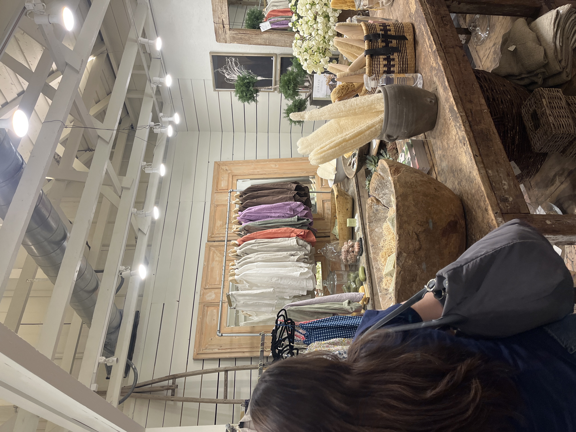 The image captures the interior of a retail store, likely a boutique, in Fredericksburg, Virginia, USA. Overhead, a white painted open ceiling structure with lights is visible. A rack of clothing hangs against the wall, displaying an assortment of garments in various colors and styles. A wooden frame surrounds the clothing rack. To the right, a wooden table displays an array of merchandise, including decorative items, brushes, and natural sponges. A person with long, brown hair, wearing a blue jacket and carrying a gray bag, stands in the foreground, appearing to be browsing or examining the items on display. The overall atmosphere suggests a curated, boutique shopping experience.