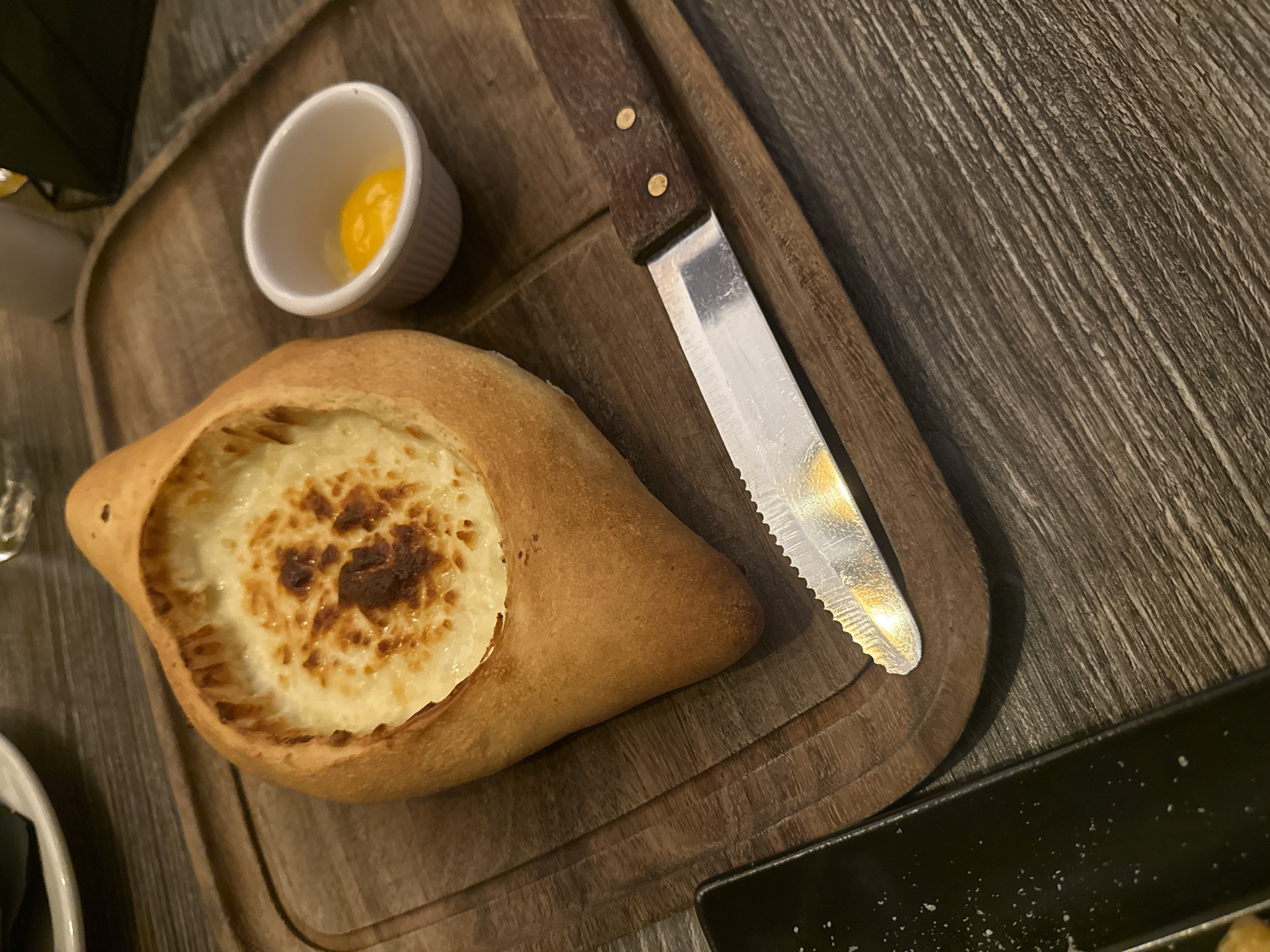 The image shows a Khachapuri, a traditional Georgian cheese-filled bread, served on a wooden board alongside a small white ramekin containing what appears to be an egg yolk and a knife. The Khachapuri is shaped like a boat with a filling of melted cheese, some parts of which are browned. The knife has a serrated blade and a wooden handle secured with rivets. The wooden board has rounded corners and edges. Part of a black plate is visible in the lower right corner. The table surface under the board has a textured, dark gray appearance, resembling wood grain. The location is implied to be Glenview, United States of America, based on instructions, but there are no specific visual cues to confirm this. There are no people visible in the image.