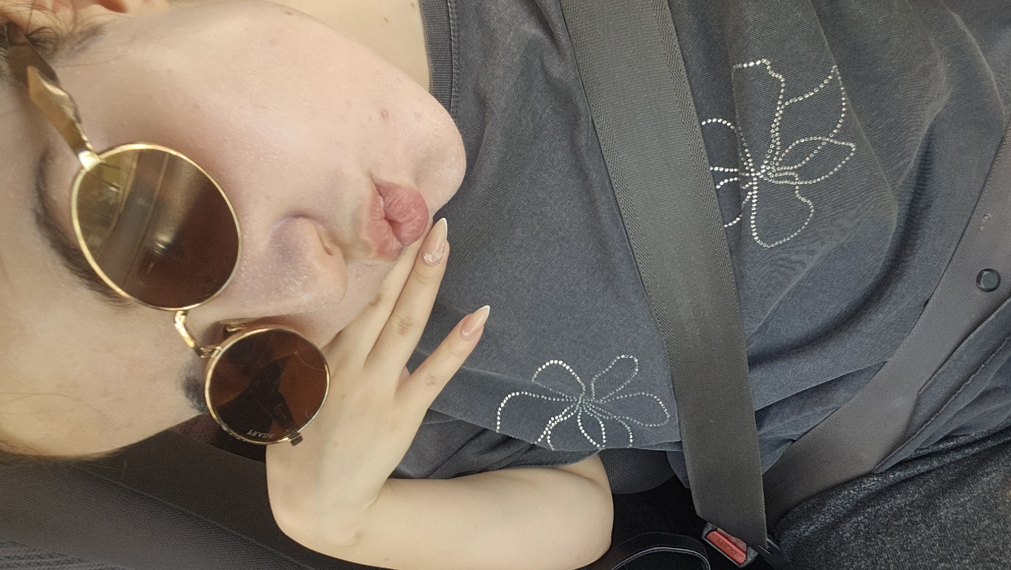 The image depicts a close-up, high-angle view of a person seated in the interior of a vehicle in Novakovo, Bulgaria. The individual, from the upper torso up, is looking towards the camera, making a pursed-lip or 'kissing' expression. They are wearing round, gold-framed sunglasses with dark lenses; the brand 'AGLET' is visible in reverse on the left lens. The person has light-toned skin with visible texture, some small blemishes on the chin, and appears to have a white, powdery residue around the nose, mouth, and chin area. Their right hand, adorned with long, light-colored almond-shaped nails (one featuring a small clear embellishment on the ring finger), is resting against their right cheek/jawline. They are wearing a dark grey t-shirt featuring two outlined floral designs made of small, reflective studs or rhinestones – one prominent on the upper left chest, another partially visible lower on the right. A dark-colored seatbelt crosses diagonally across the person's chest, with its red release button visible to the far left. The background consists of the dark, indistinct interior of the vehicle.