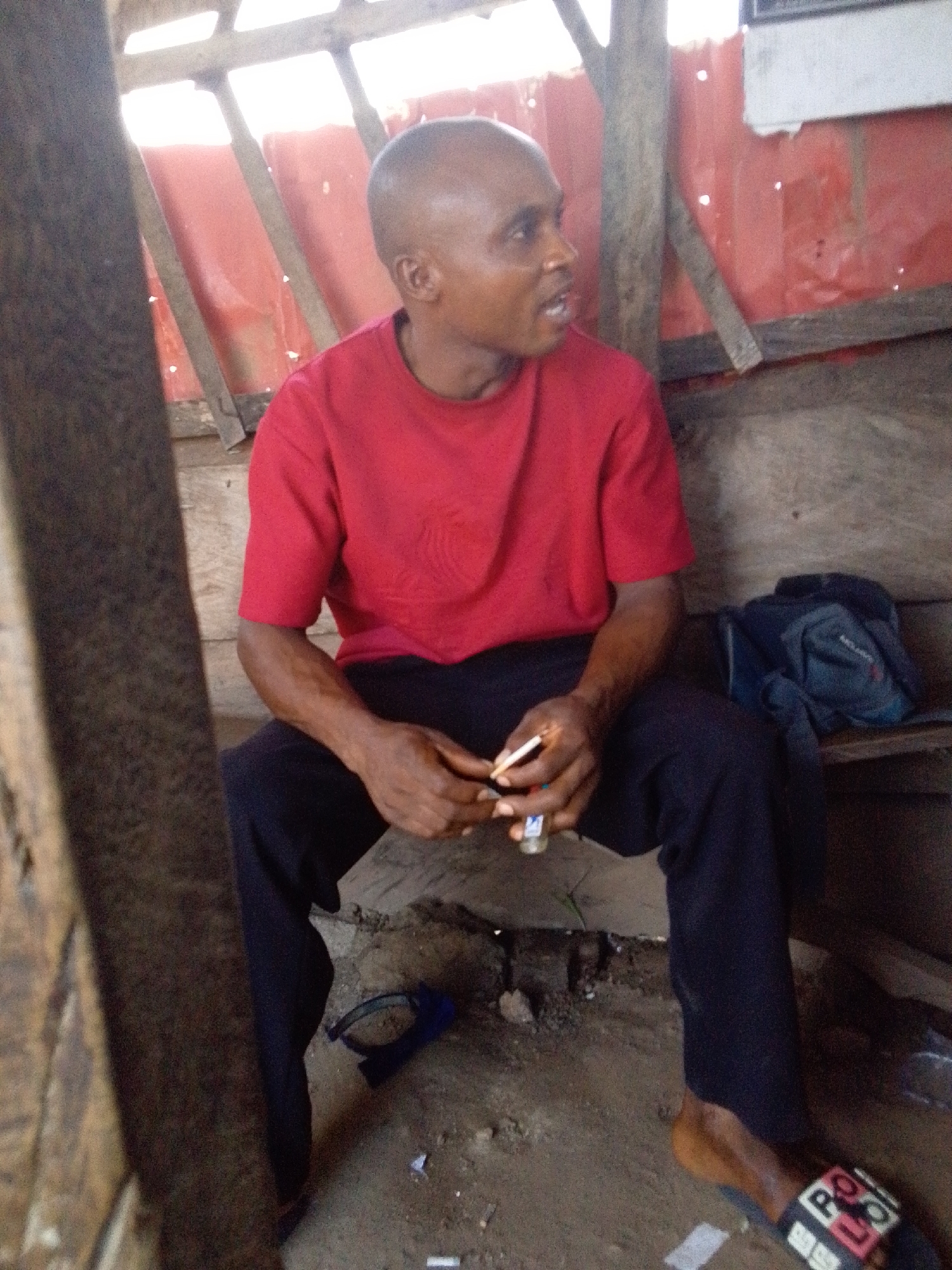 An adult man with a shaved head, wearing a red short-sleeved t-shirt and dark trousers, is seated on a wooden bench or low platform within a rustic structure. He is looking towards the right side of the frame with his mouth slightly open, appearing to be speaking or reacting to something off-camera.
In his right hand, he holds a cigarette, and in his left hand, he holds a small, possibly blue and white, lighter, suggesting he is in the process of lighting or has just lit the cigarette. On his right foot, he wears a black and white slide sandal with the word "POLO" clearly visible in white letters. His left foot is either bare or obscured.
The setting appears to be a simple, semi-enclosed space, possibly a shack or shelter. The background features red corrugated material, which could be a metal sheet or tarp, forming part of a wall or roof. Wooden beams and supports are visible, framing the background. A dark blue backpack or bag rests on the wooden surface next to him. The ground beneath him is dirt or concrete with some scattered debris, and a blue object (possibly a strap or fallen item) is also on the ground. The lighting suggests it is daytime.