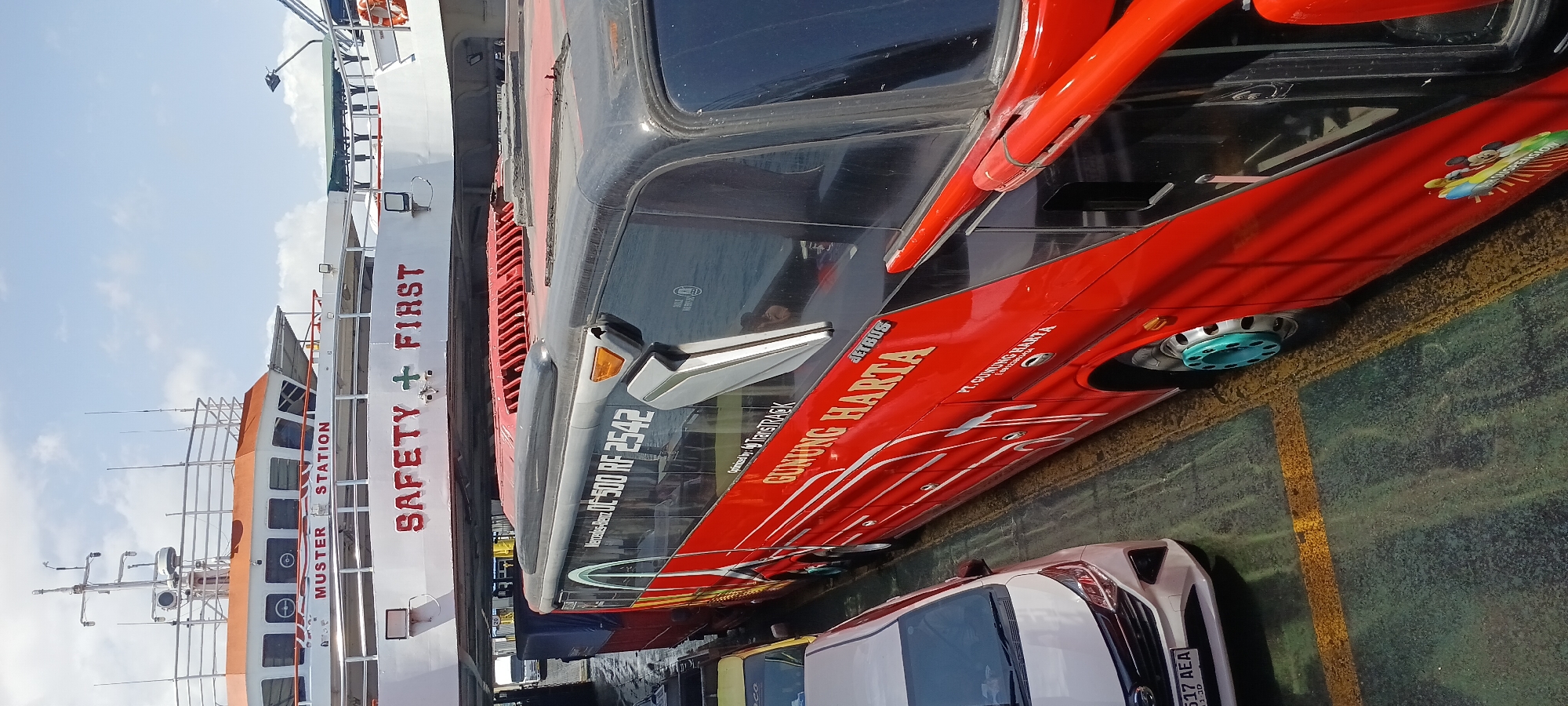 The image captures a bright, sunny day with a clear blue sky and some white clouds. The foreground features the side of a red and black bus, possibly a "Gunung Harta" bus, on a ferry deck. The bus has "JETBUS" written on its side and a license plate is partially visible on a white car parked next to it, reading "517 AEA". The bus is positioned parallel to the ferry's edge. In the background, a white ferry with "MUSTER STATION" and "SAFETY FIRST" written on its side is visible. The ferry deck appears to be made of weathered green and yellow painted concrete, with visible tire marks. The overall scene suggests vehicles being transported on a ferry, likely in the coastal area of Melaya, Indonesia, given the "Gunung Harta" bus branding, which is a prominent bus operator in Bali and Java, Indonesia.
