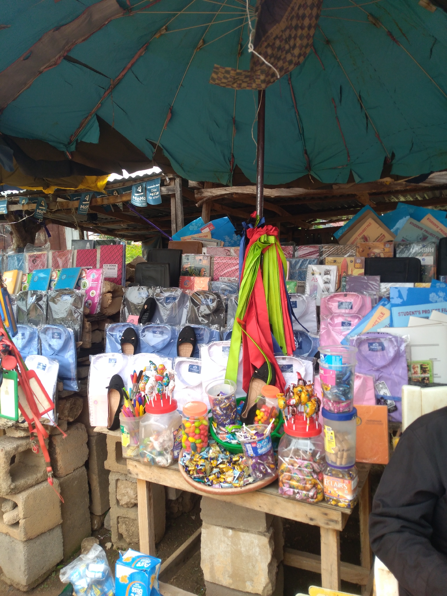 This is a medium shot photograph of a street market stall in Jos, Nigeria. The stall is covered by a tattered turquoise tarp, with some corrugated metal roofing visible underneath. The stall is packed with a variety of goods, including stacks of men's shirts in various colors (white, light blue, pink, and purple), neatly folded and packaged in plastic. There are also notebooks, folders, and other stationery items displayed.
In the foreground, a wooden table holds an assortment of candies and lollipops in clear plastic containers and bowls. A pair of black high-heeled shoes and a pair of black slip-on shoes are placed on top of some folded shirts. Several colorful ribbons are draped over a central pole at the stall, adding a festive touch. The stall is built against a backdrop of cinder blocks, some of which are stacked precariously. In the lower right corner, a person wearing a black garment is partially visible, with their arm resting on a stall surface. Some plastic bags of what appear to be packaged snacks are on the ground near the base of the stall. The overall scene depicts a busy, open-air market environment.