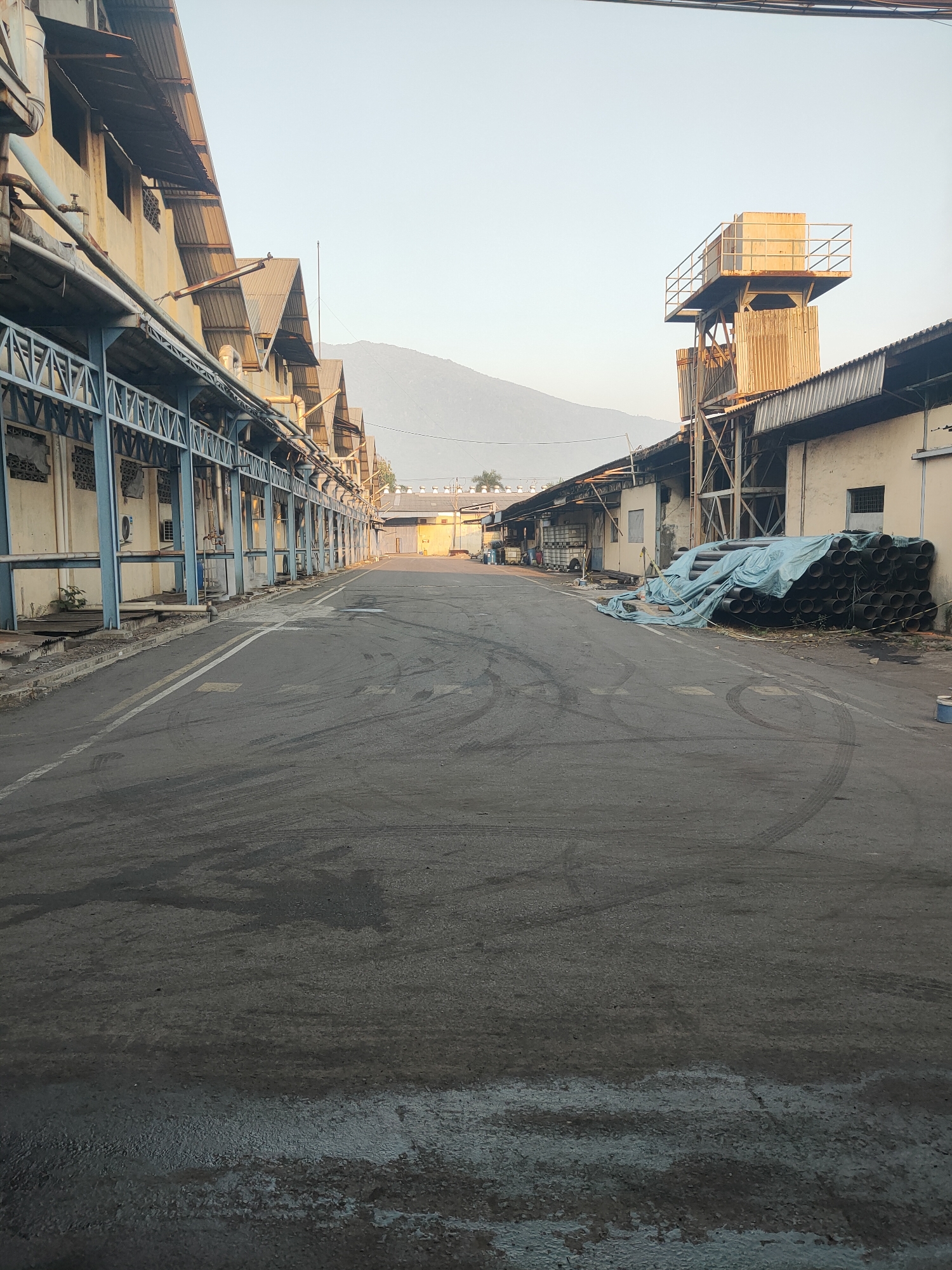 An outdoor, medium-shot, eye-level photograph captures a view down a paved industrial road. The road is flanked by buildings on both sides, with a mountain range visible in the background under a clear, pale sky.
On the left, a long, low building with a corrugated metal roof extends into the distance. Its facade is light-colored, and a blue metal structure runs along its front, creating a covered walkway. Pipes and conduits are visible along the building's exterior.
On the right, a taller structure, possibly a water tower, rises above a single-story building. This building also has a corrugated metal roof. In front of this building, a large pile of black pipes, covered with a light blue tarp, is stacked on the ground.
The asphalt road shows tire marks and wet patches, suggesting recent activity or rainfall. White lines mark the center of the road. The overall impression is that of a functional but somewhat aged industrial area. The location is identified as a facility in Beji, Indonesia. There are no people visible in the photograph.