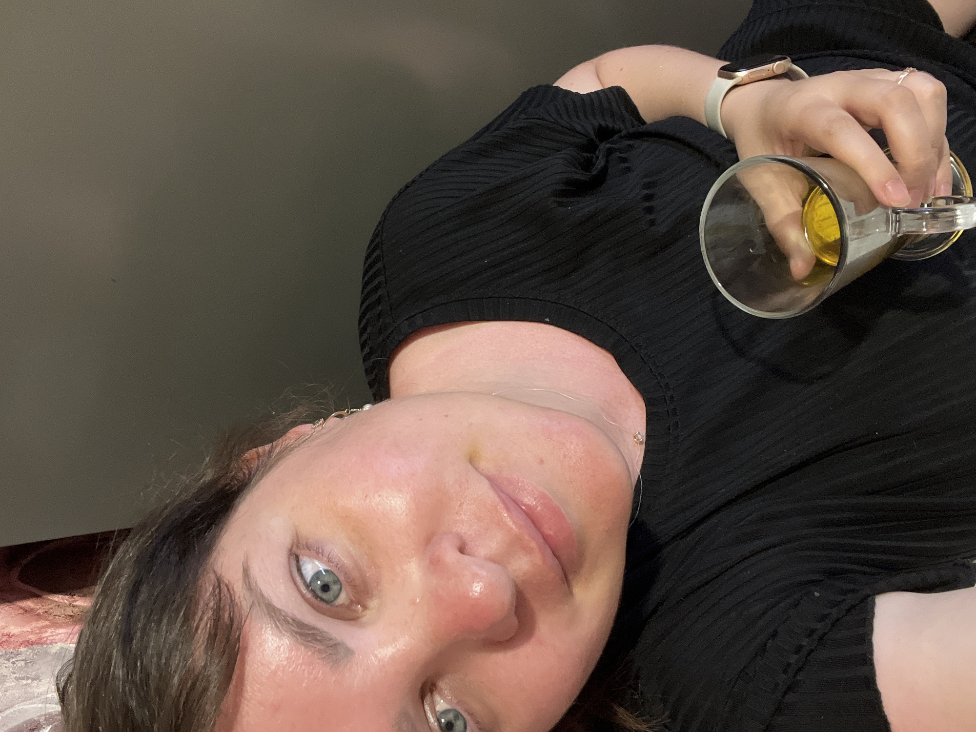This is a close-up, selfie-style photograph of a woman with light skin and blue-green eyes, taken from a high angle. She is wearing a black, ribbed t-shirt and a light-colored smartwatch on her left wrist. A thin, delicate necklace with a small pendant is visible around her neck. She is holding a clear glass with a handle, containing a yellow liquid, in her right hand. The background is a dark, plain wall. The lighting suggests an indoor environment, possibly a home. There is no discernible urban or natural landscape visible in the image, therefore the location in Noviy, Russia cannot be determined.