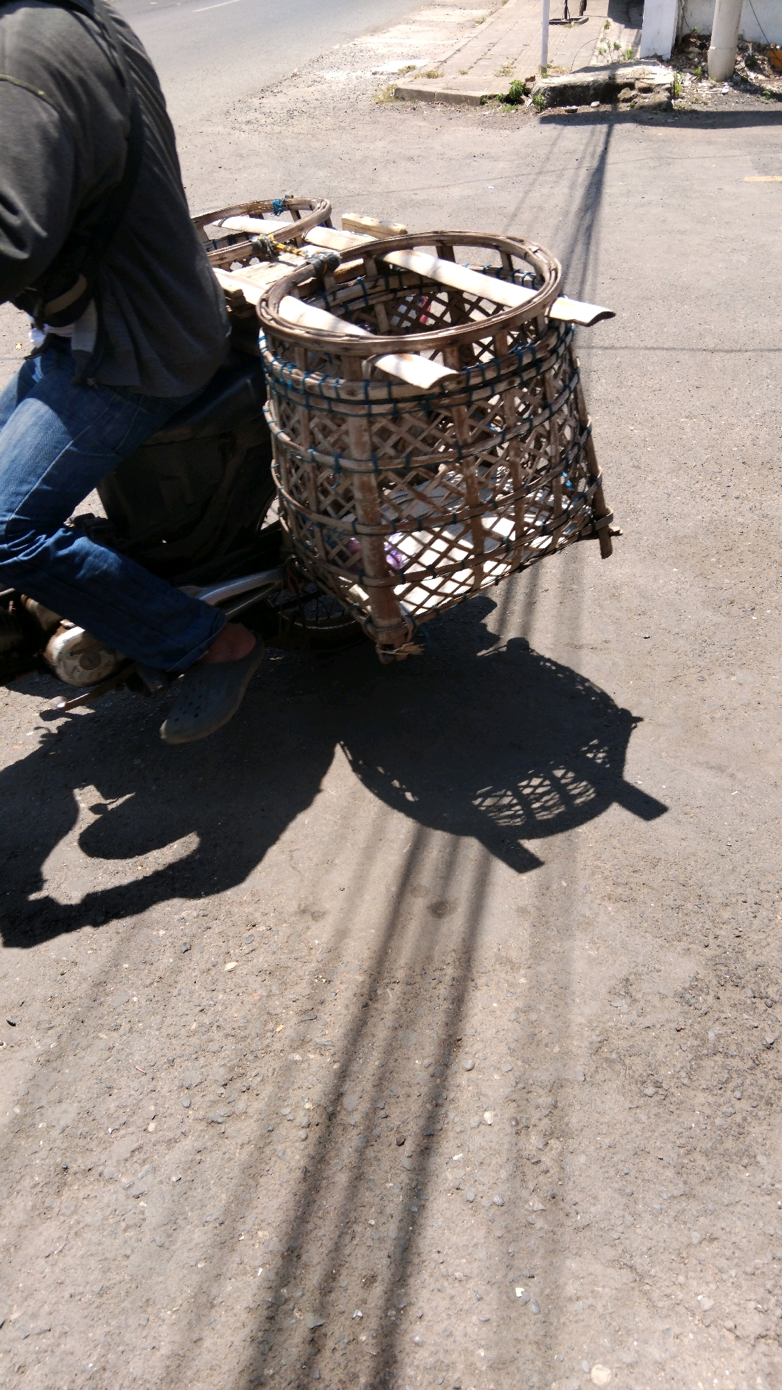 A person is sitting on a motorbike with two woven baskets attached to the back. The person is wearing a dark jacket and blue jeans, and the right foot, clad in a dark grey clog, is resting on the ground. The motorbike has a dark frame and a black bag behind the seat. The baskets are made of woven bamboo and appear to be empty or lightly filled with white material. The scene is set on a sun-drenched, rough asphalt road in Watesalit, Indonesia. The shadow of the motorbike and baskets, as well as overhead power lines, stretch across the road, indicating a bright sunny day. In the background, a concrete sidewalk and a low wall can be seen, suggesting a semi-urban or roadside environment. The overall impression is one of an everyday scene of transportation and possibly commerce in a tropical setting.