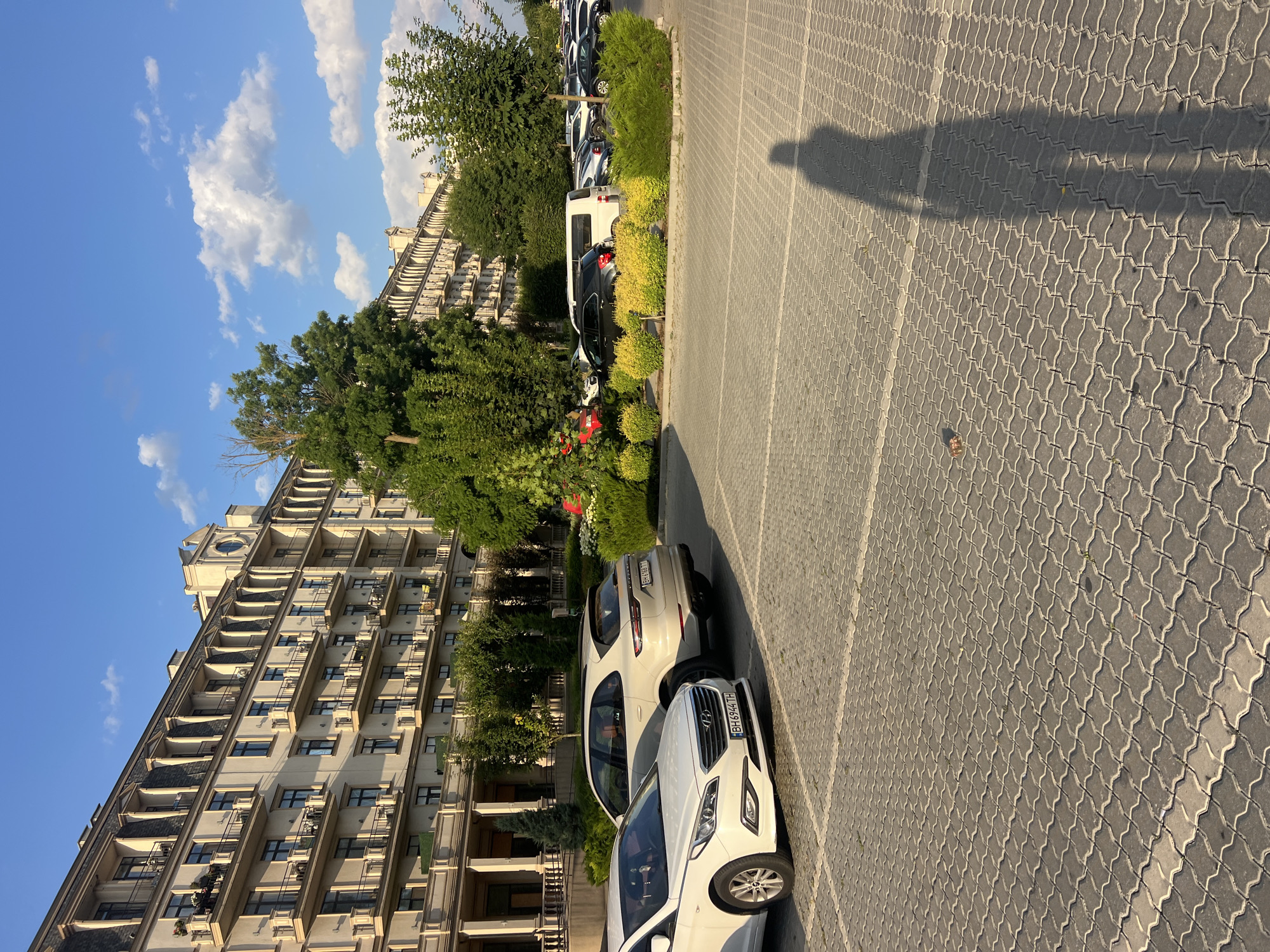 An outdoor, daytime, low-angle shot captures a parking lot in front of a large, multi-story building. The building, with its beige facade, numerous balconies, and ornate architectural details, appears to be an apartment complex or hotel. A line of trees and manicured bushes with yellow and green foliage separates the parking lot from the building. Several cars are parked in the lot, including a white sedan in the foreground and a cream-colored SUV behind it. The parking lot is paved with gray interlocking bricks. The sky is blue with scattered white clouds, and a long shadow of a person is cast across the pavement, suggesting a sunny day. The scene is located in Fontanka, Ukraine.
