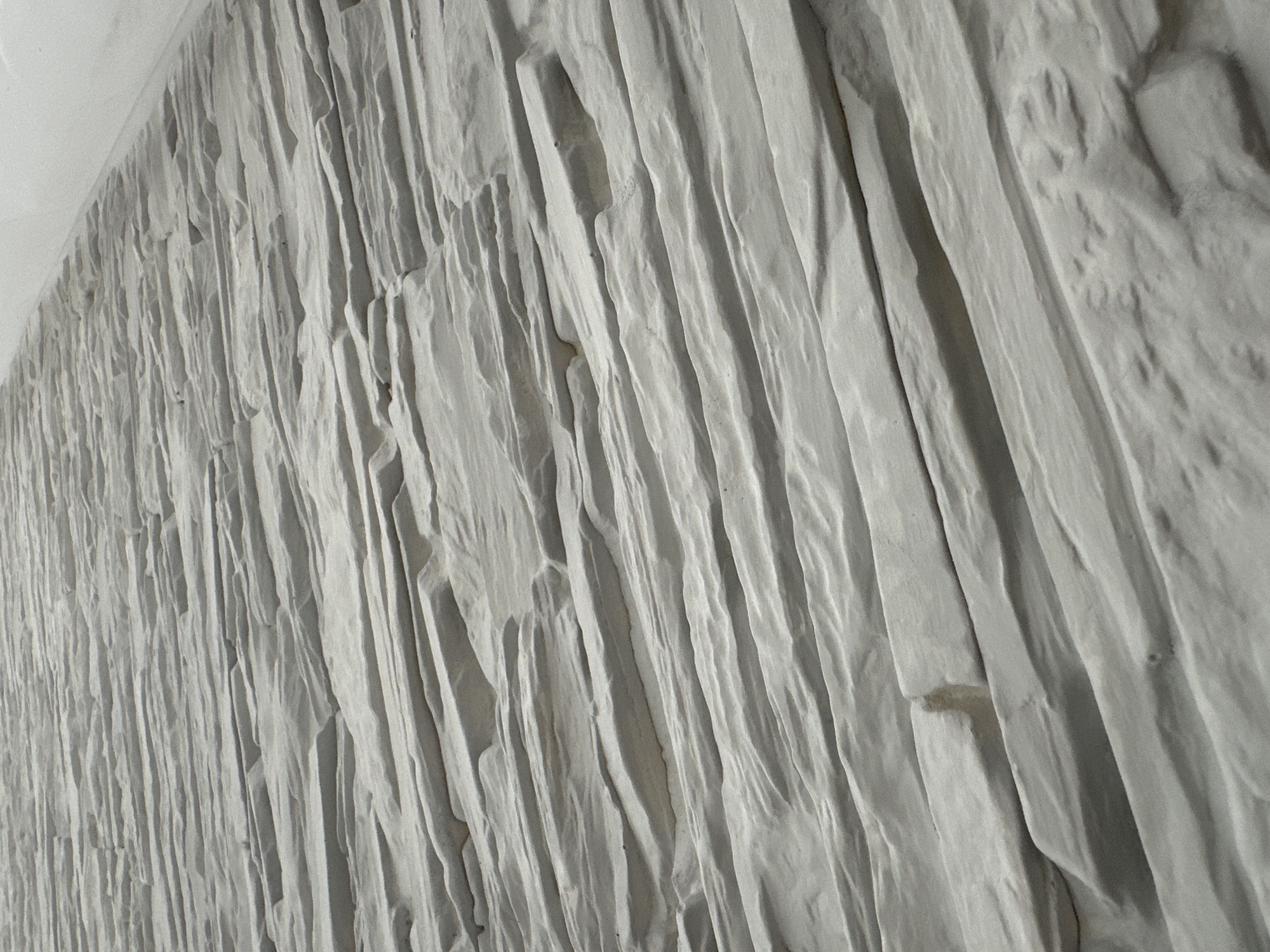 The image displays a textured wall or architectural surface, captured from an oblique, upward-looking perspective. The dominant subject is a relief pattern characterized by prominent, irregular, and predominantly vertical ridges and valleys. The surface is rendered in shades of light gray and off-white, with the interplay of light and shadow strongly emphasizing its three-dimensional quality and depth.
The texture suggests natural rock formations, possibly resembling sedimentary layers or a roughly chiseled stone wall. It features a mix of smoother, elongated forms and more rugged, undulating sections. These vertical lines create a dynamic visual flow across the surface. The perspective allows the texture to recede into the distance on the left side of the frame, where it becomes less defined. In the upper-left corner, a flatter, smoother light-colored surface, likely a ceiling or an adjoining wall, forms a distinct boundary.
There is no discernible activity or event taking place; the image is a static portrayal of a decorative or structural element. The lighting suggests an indoor setting with illumination designed to highlight the surface's tactile qualities. A small, light brown smudge or discoloration is visible on one of the ridges in the closer view. No text is visible in the image.