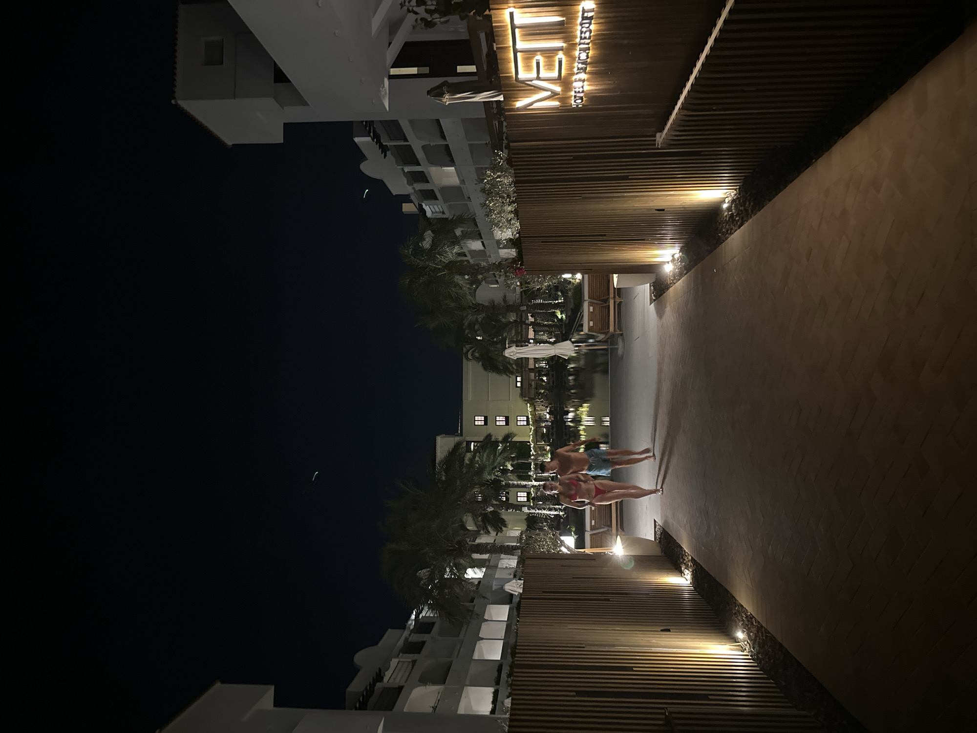 An exterior night shot captures a couple walking on a pathway towards a body of water. The pathway is lined with wooden slats and illuminated by warm lights. To the right, a sign reads "METT HOTEL & BEACH RESORT." Palm trees and white buildings with illuminated windows flank the scene, suggesting a resort or hotel environment in Playa Del Sol-villacana, Spain. The sky is dark and clear, with a faint streak of light visible in the upper portion of the image, possibly an airplane or satellite. The couple is walking side-by-side, appearing to be enjoying a relaxed evening stroll. The overall atmosphere is serene and inviting.