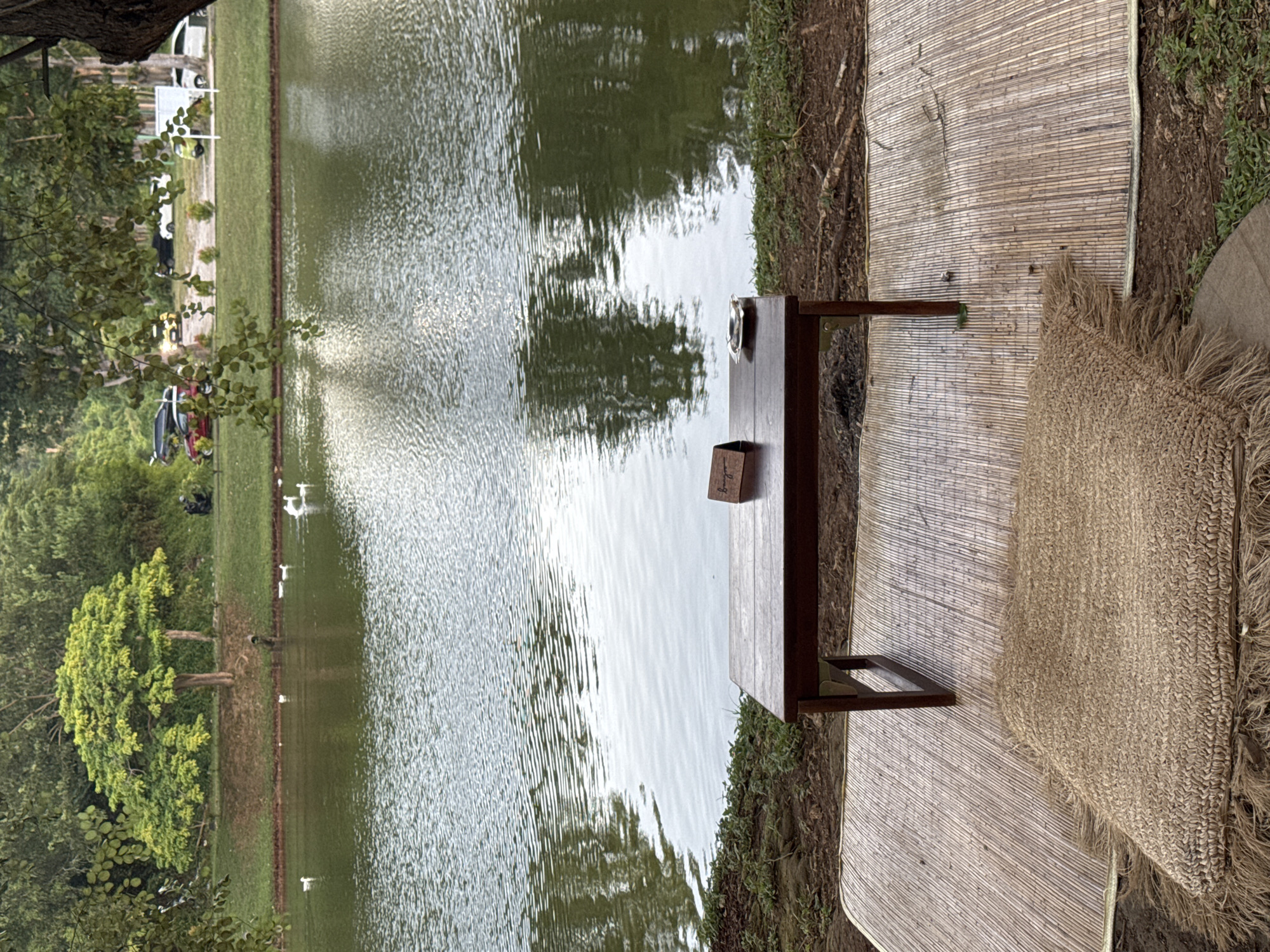 The image shows a lakeside scene in Pecatu, Indonesia. A narrow wooden table sits on a woven mat next to the lake's edge. There's a small box and a metal object (likely a cup or saucer) on the table. A straw mat is placed next to the woven mat, and trees line the opposite bank of the lake. The water shows reflections of the sky and trees. There's grass on the bank between the mat and the water.
