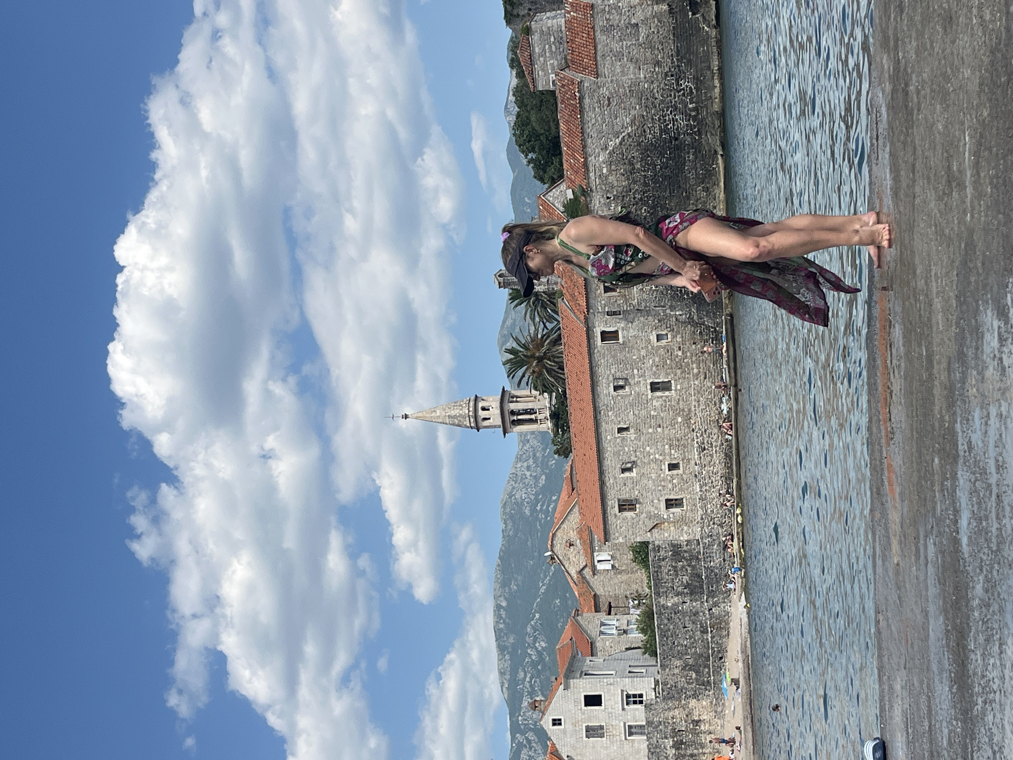 A woman stands on a stone pier, looking out at the water. She is wearing a colorful sarong and a dark visor. Behind her, the ancient walled city of Budva, Montenegro, is visible. The city features stone buildings with red tile roofs and a prominent bell tower. The Adriatic Sea stretches out before the city, and in the distance, mountains rise under a sky filled with white clouds. People can be seen on the small beach and swimming in the water in front of the city walls. The scene suggests a sunny day in a historic coastal town.