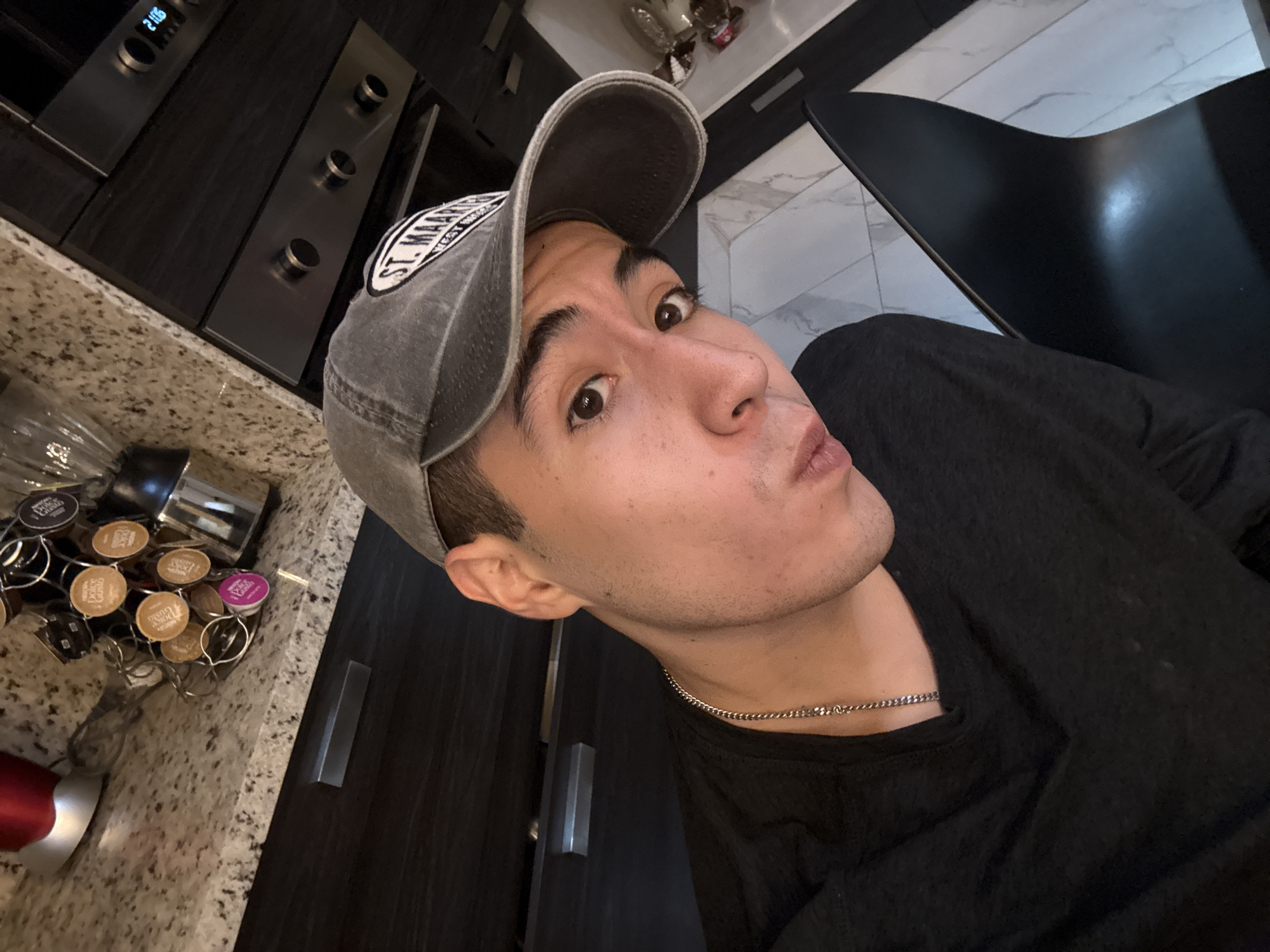 A selfie of a person with light skin wearing a grey baseball cap with a patch that reads "St. Mary's" and a black t-shirt, taken indoors. The background includes a dark kitchen cabinet with a built-in oven, a countertop with coffee capsules in a metal holder, and a black chair. The location is not San Bernardino Tlaxcalancingo, Mexico.
