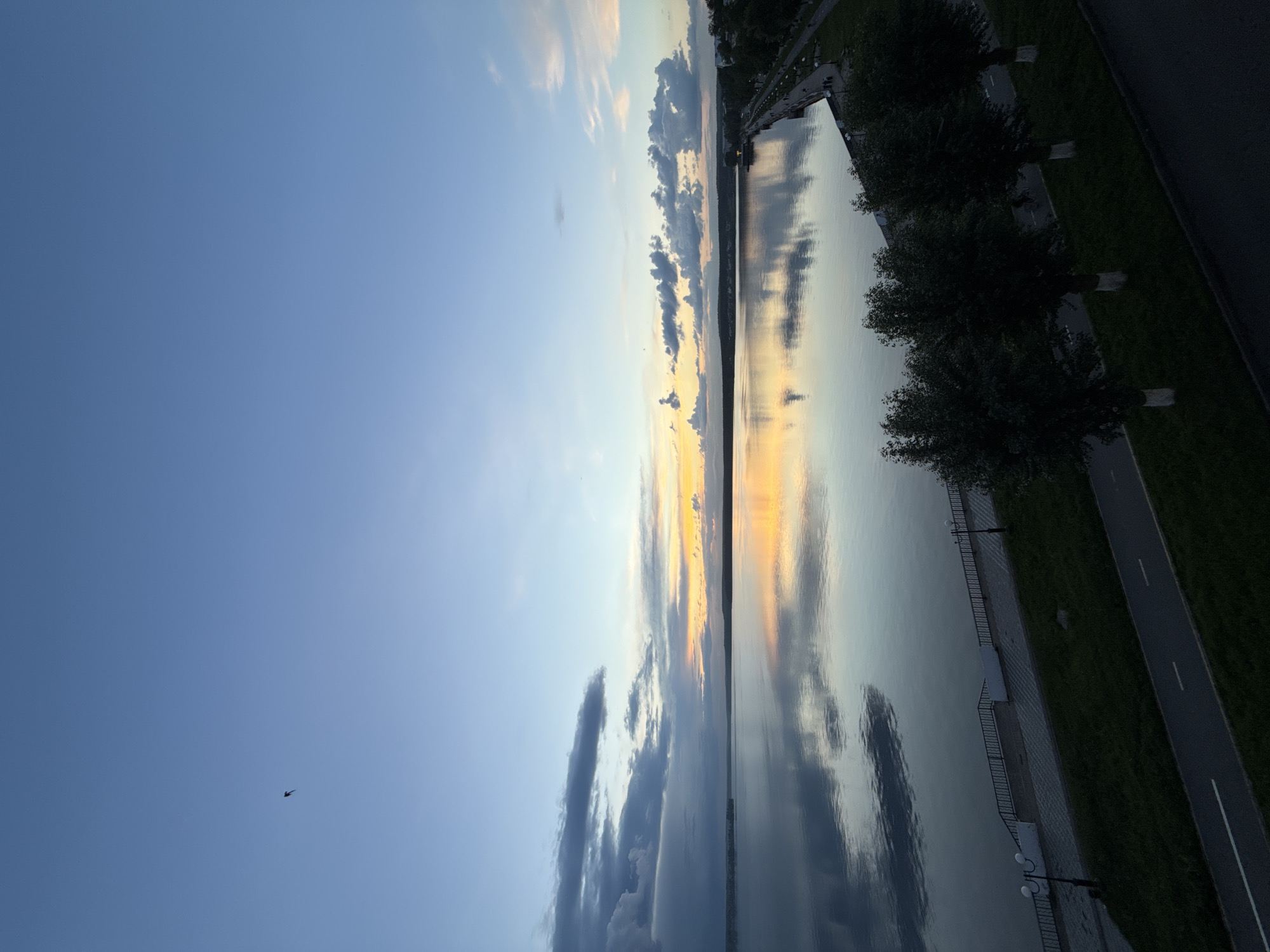 The image captures a wide view of the Votkinsk Reservoir in Votkinsk, Russia, during what appears to be twilight. The sky is a gradient of blues, with hints of yellow and orange near the horizon, suggesting either sunrise or sunset. Scattered clouds are visible, some dark and heavy on the left, and lighter, more wispy clouds on the right.
The calm water of the reservoir perfectly reflects the sky and the distant shoreline, creating a mirror-like effect. The shoreline features a promenade or embankment with a railing, lined with trees and lampposts. A paved path, possibly a sidewalk or bike lane, runs along the grassy area in the foreground. Four trees with whitewashed trunks are prominently featured in the lower right quadrant of the image. A single bird is captured in flight in the upper left portion of the sky. The overall impression is one of tranquility and natural beauty.
