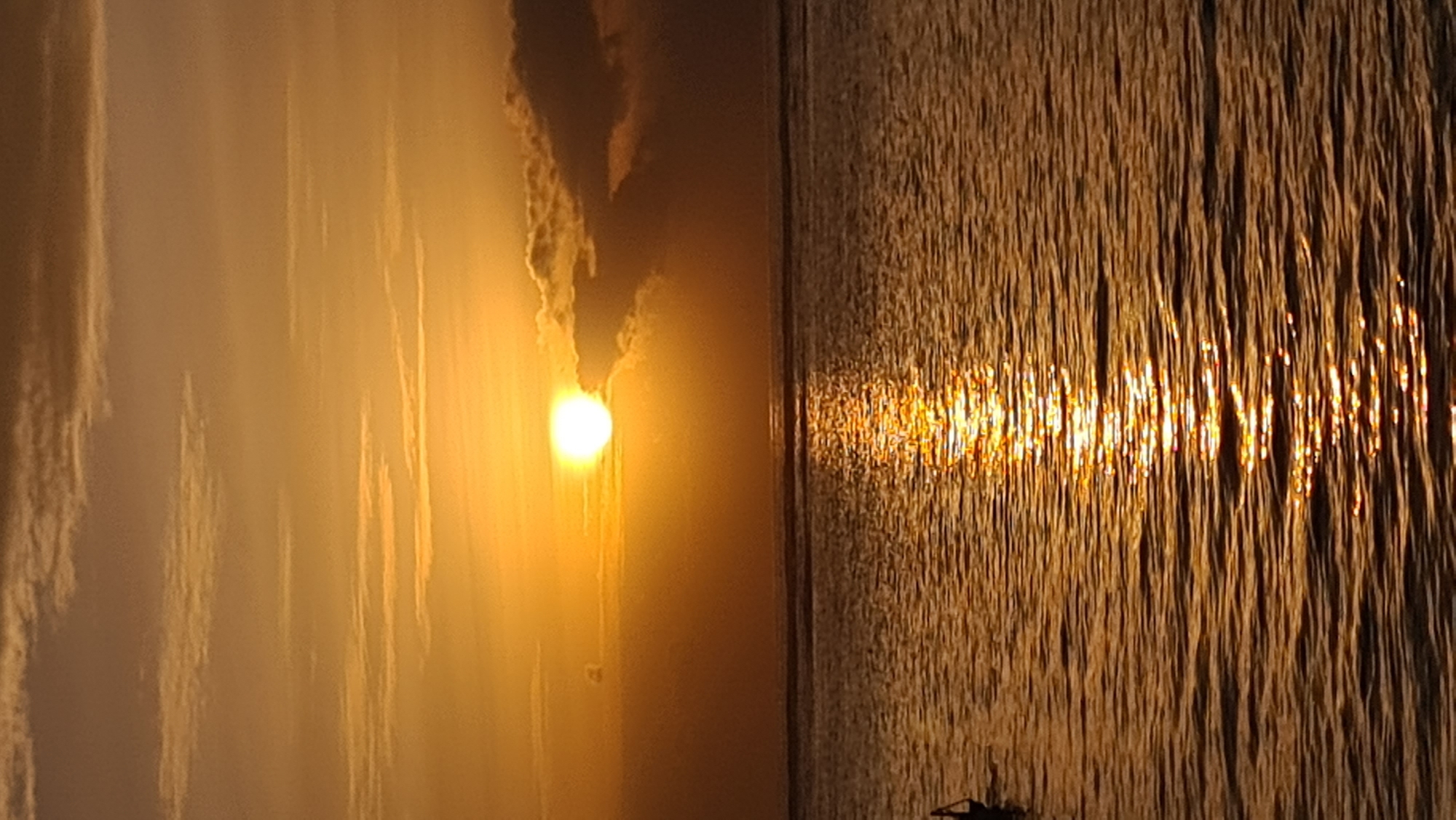 A sunset over the ocean in Denpasar, Indonesia, casts a warm, golden glow across the sky and water. The sun, a bright, white orb, is partially obscured by dark clouds on the right side of the frame, creating a dramatic contrast. Wispy, light-colored clouds are scattered across the upper portion of the sky, adding texture to the atmospheric scene.
The ocean surface is rippled, reflecting the intense orange and yellow hues of the sunset, creating a shimmering pathway of light that stretches towards the viewer. The horizon line is clearly defined, separating the sky from the water. On the far left of the image, a small, dark silhouette of what appears to be a person in a boat or on a buoy is visible in the water. The overall scene evokes a sense of tranquility and natural beauty, characteristic of a tropical sunset.