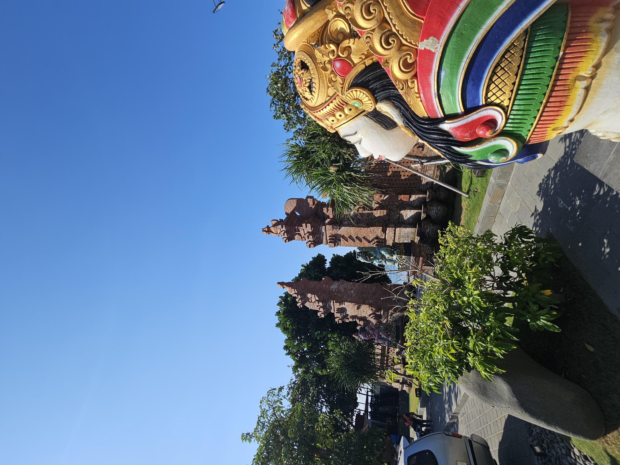 The image depicts a section of the Bajra Sandhi Monument in Denpasar, Bali, Indonesia. In the foreground, a large, vibrantly colored statue of a Balinese woman is positioned to the right, her head and upper torso filling the frame. Her intricate headdress is gilded with red and green accents, and her attire is a colorful display of red, green, blue, and yellow patterns. Behind the statue, a paved walkway leads towards a pair of ornate, brick gateways, characteristic of Balinese temple architecture. Green foliage and palm trees are visible between the gateways and in the background. To the left, a silver car is parked, and several people are seated in the shade of a tree. The sky above is a clear, bright blue, indicating a sunny day. The scene conveys a sense of cultural heritage and public space.