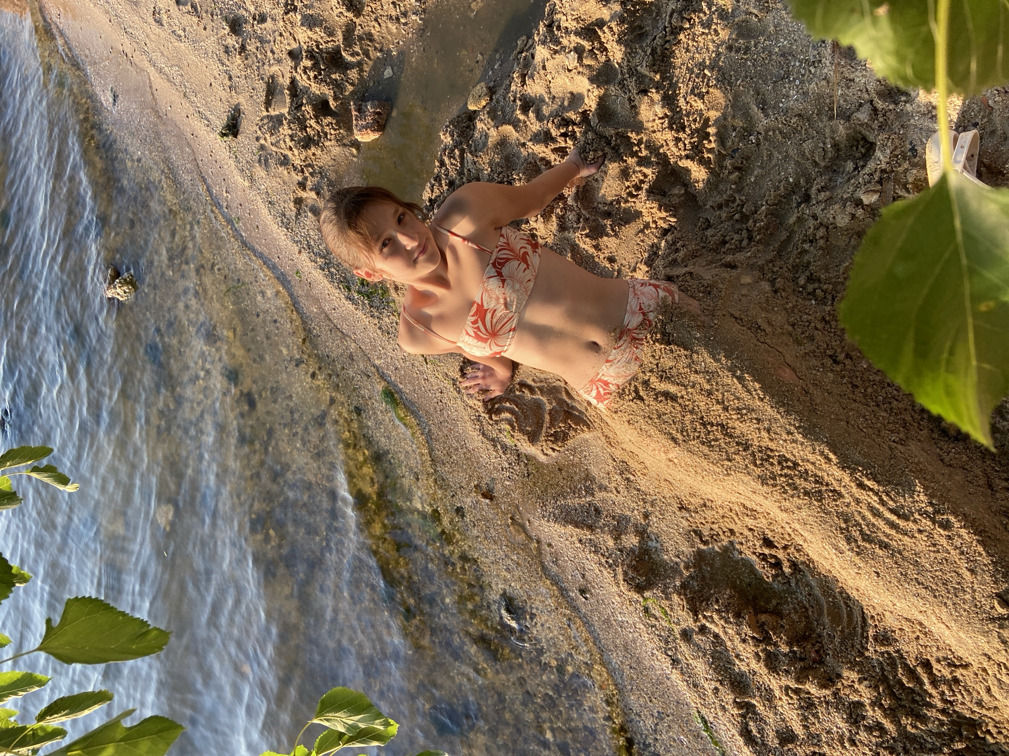 A young person is lying on a sandy beach next to the water. They are wearing a bikini with a coral and white floral pattern. The person is partially buried in the sand, with their body angled towards the water. Their left arm is extended, and their right arm is bent, with their hand resting on the sand. Their head is turned towards the camera, and they are looking directly at the viewer with a slight smile. The sunlight casts a warm glow on the scene, suggesting it is either morning or late afternoon. The water is calm and reflects the sunlight. Green leaves from a nearby plant are visible in the foreground, partially obscuring the view. The sand is wet near the water's edge, indicating that the tide may be coming in or going out. The overall mood of the image is relaxed and playful.
