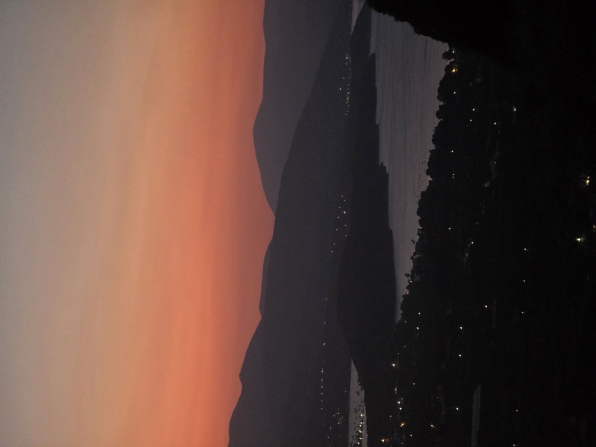 The image captures a twilight view of the Municipal Unit of Asini, Greece. The sky is a gradient of warm orange and lighter hues, transitioning into a soft grey at the top. Silhouetted mountain ranges dominate the background, with layers of dark, indistinct shapes. In the middle ground, a body of water, likely a bay or sea, is visible, reflecting the dim light. Along the coastline and nestled within the hills, numerous small lights twinkle, indicating a populated area, possibly the town of Asini or nearby villages. The foreground is heavily shadowed, with the dark forms of trees or a hillside obscuring much of the detail. The overall impression is one of peaceful evening scenery.