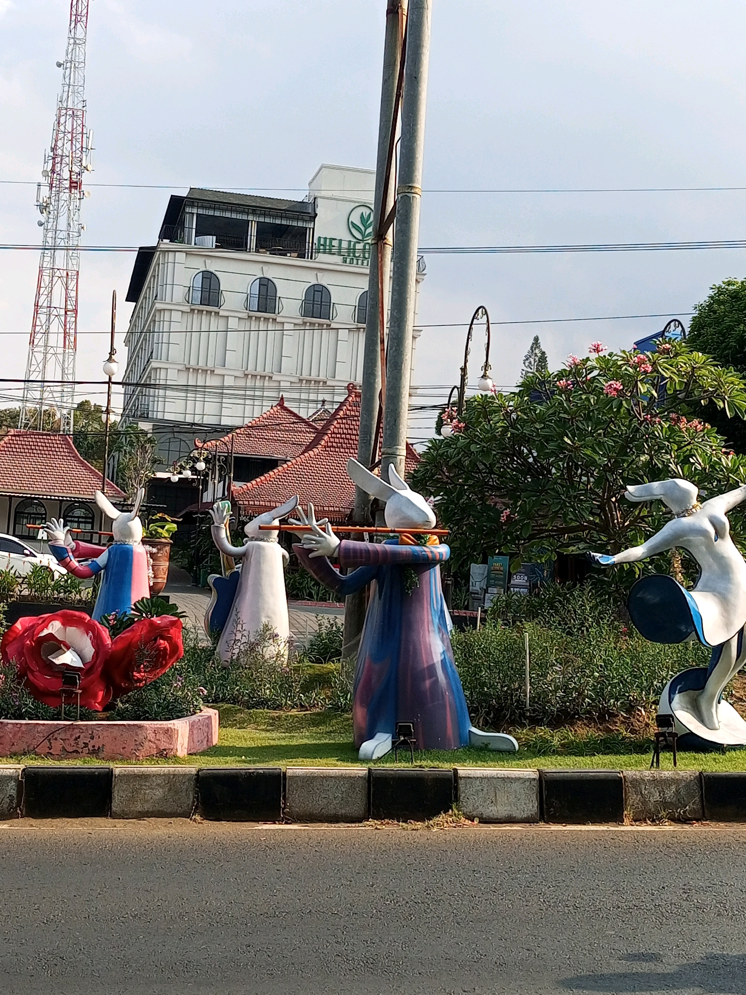 The image captures a street scene in Batu, Indonesia, featuring a collection of rabbit-themed sculptures arranged in a landscaped area. In the foreground, a paved road is visible, bordered by a concrete and black curb. Behind the curb, a garden bed contains grass, low-lying plants, and several large, stylized red roses. The main subjects are white rabbit figures with elongated ears, dressed in colorful, flowing robes. Three of the rabbits are depicted playing musical instruments, resembling flutes, and appear to be in a synchronized performance. A fourth rabbit, also white, is sculpted in a dynamic, dancing pose.
In the background, a tall white building with arched windows and a dark-tiled roof dominates the upper portion of the scene. A large communication tower, red and white, stands prominently to the left of the building. Several power lines crisscross the sky. Mature trees with pink flowers are visible behind the sculptures, adding to the lushness of the environment. The overall impression is one of an artistic display intended to attract attention in a public space, likely a tourist attraction or a commercial area in Batu, known for its gardens and natural beauty.