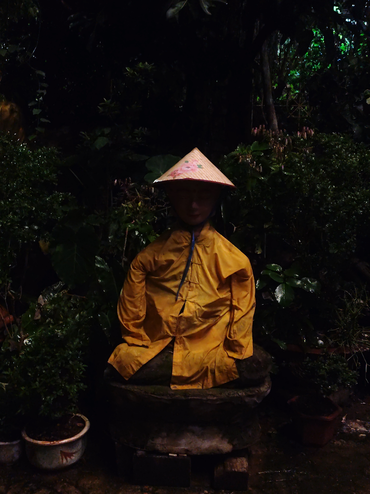 An outdoor, low-light photograph captures a statue or figure adorned in traditional Vietnamese attire, set amidst dense foliage. The central subject is a figure, possibly a statue, positioned in a sitting or squatting posture on a dark, rounded stone base. It wears a bright yellow, long-sleeved raincoat, which appears somewhat wrinkled, and a classic conical hat (nón lá) with subtle floral patterns in pink and white. The face of the figure is largely obscured by the shadow cast by the hat and the overall dim lighting.
The setting is a lush, verdant environment, likely a garden or a natural area, consistent with the location context of Da Lat, Vietnam. The background is dominated by dark green leaves, shrubs, and tree trunks, suggesting a dense growth. Several potted plants are visible at the bottom left of the frame, contributing to the garden aesthetic. Some leaves appear glossy, hinting at a damp environment or recent rain, which is reinforced by the presence of a raincoat on the figure.
The image is captured in very low light conditions, indicating either nighttime or a heavily shaded area. There are deep shadows throughout, with only the yellow raincoat and parts of the conical hat being distinctly illuminated. The overall mood is serene and somewhat mysterious due to the darkness and the static nature of the figure. No explicit activity is taking place, and no visible text is present in the image.