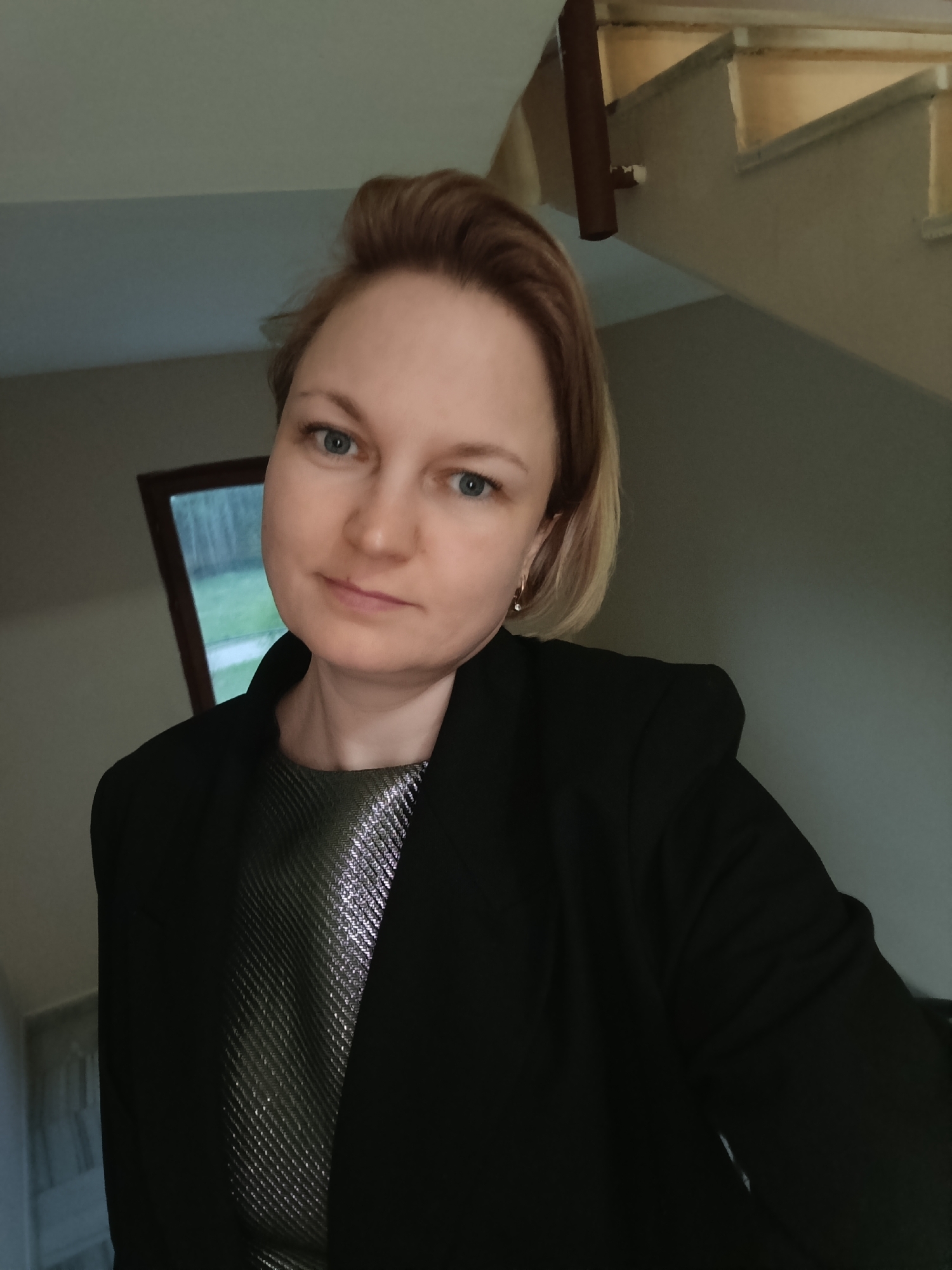 A woman is posing for a selfie indoors. She has short, blonde hair and blue eyes. She is wearing a black blazer over a shimmering silver and green top. In the background are stairs and a window with trees visible outside. The exact location within Марковский, Russia, cannot be determined from the image.