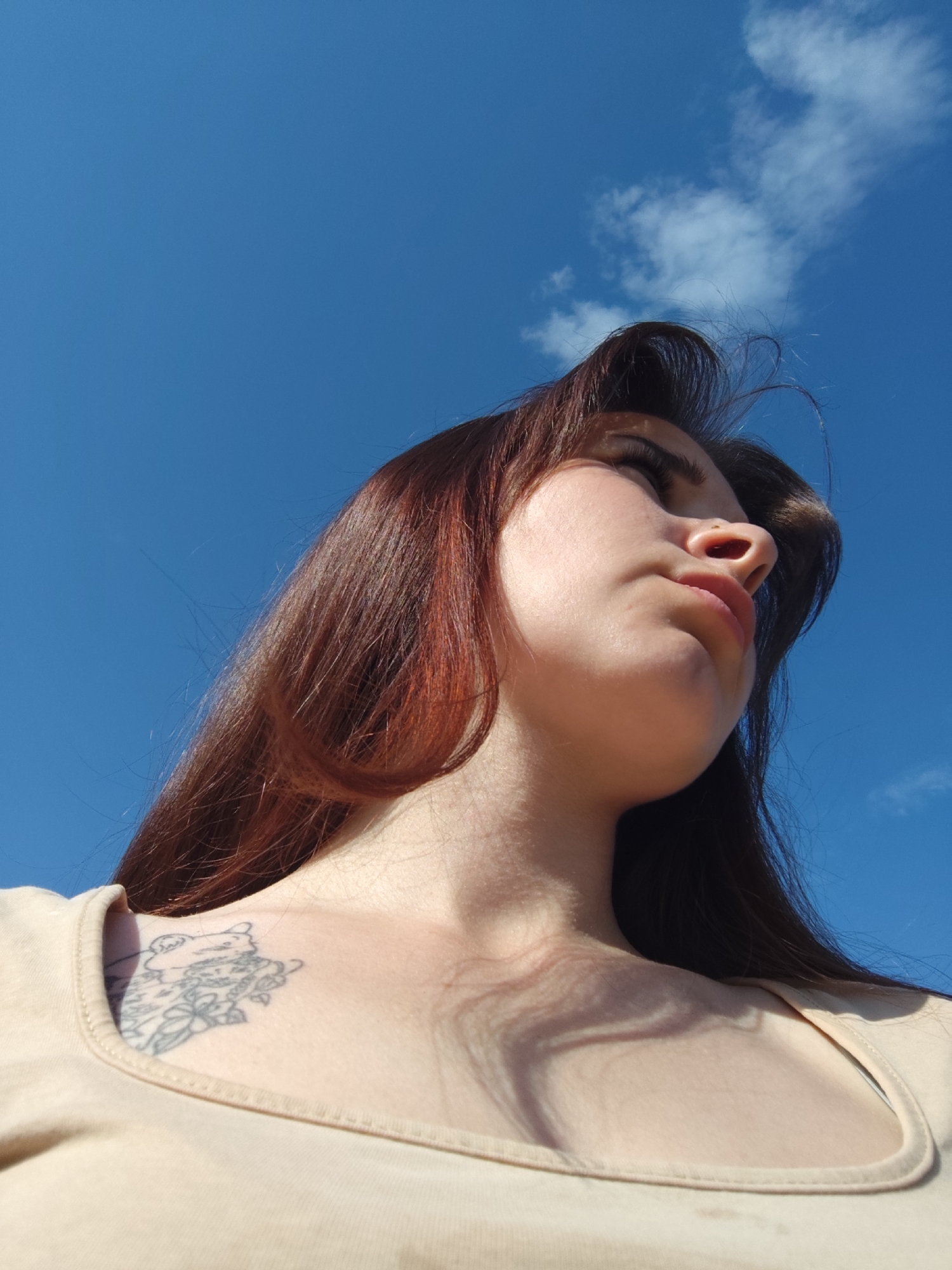 A low-angle, medium shot depicts a light-skinned woman with long, auburn hair, gazing upwards and slightly to the right. She has a nose piercing on the left side. She wears a light tan shirt with spaghetti straps. A tattoo of a bear and floral elements is visible on her left collarbone area. The background is a bright blue sky with a few white clouds in the upper right corner. The location is not discernible from the image, so it is not possible to tell where this photo was taken.