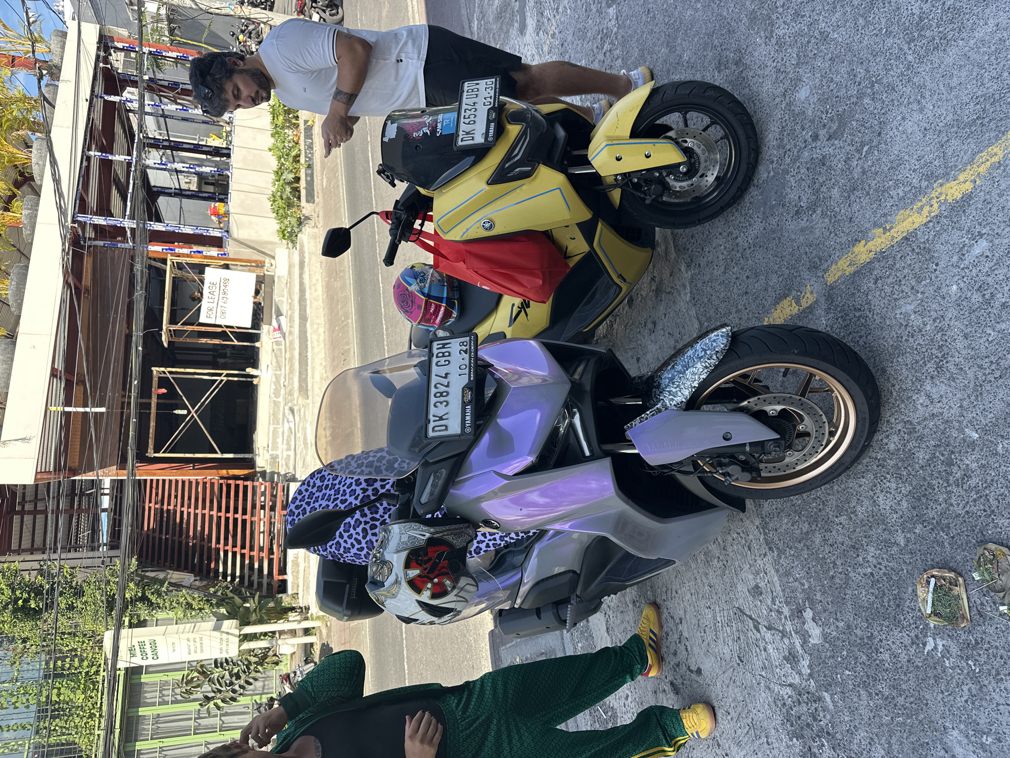 The image shows two parked Yamaha motorcycles in North Kuta, Indonesia. The foreground motorcycle is lavender and gray, with a blue and white leopard-print cloth on the front. A helmet with a decorative face design rests on it. The license plate reads "DK 3824 GBN". A person in a green tracksuit and yellow shoes stands near the motorcycle, with the lower portion of their body visible.
The motorcycle behind it is yellow. A red bag is on the seat, and the license plate reads "DK 6534 UBV". A man in a white shirt and black shorts is bending over the motorcycle, looking at his phone.
In the background, there is a building under construction with exposed metal framework and a "FOR LEASE" sign. Vegetation is also visible, as is a sign for "MIEL COFFEE CANGGU". The street is paved and marked with yellow lines.