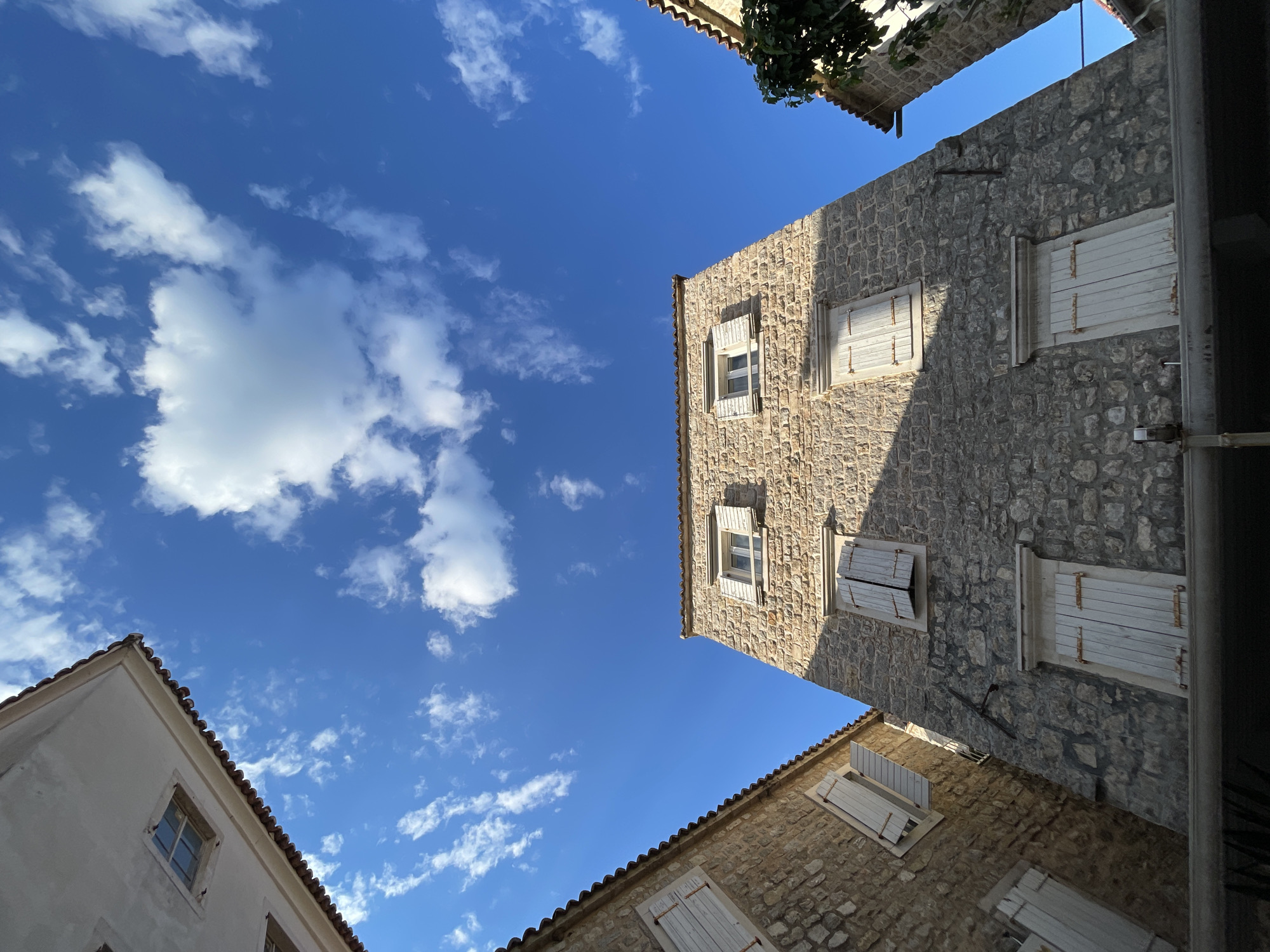 The image is a low-angle shot of buildings in an urban setting, likely a narrow street or alley. The architecture features stone buildings with white-framed windows, some of which are partially shuttered. The sky is a clear blue with scattered white clouds. A small patch of greenery is visible at the top of the frame, possibly from a tree or vine growing on a building. The location appears to be in the city of Unknown, Unknown.