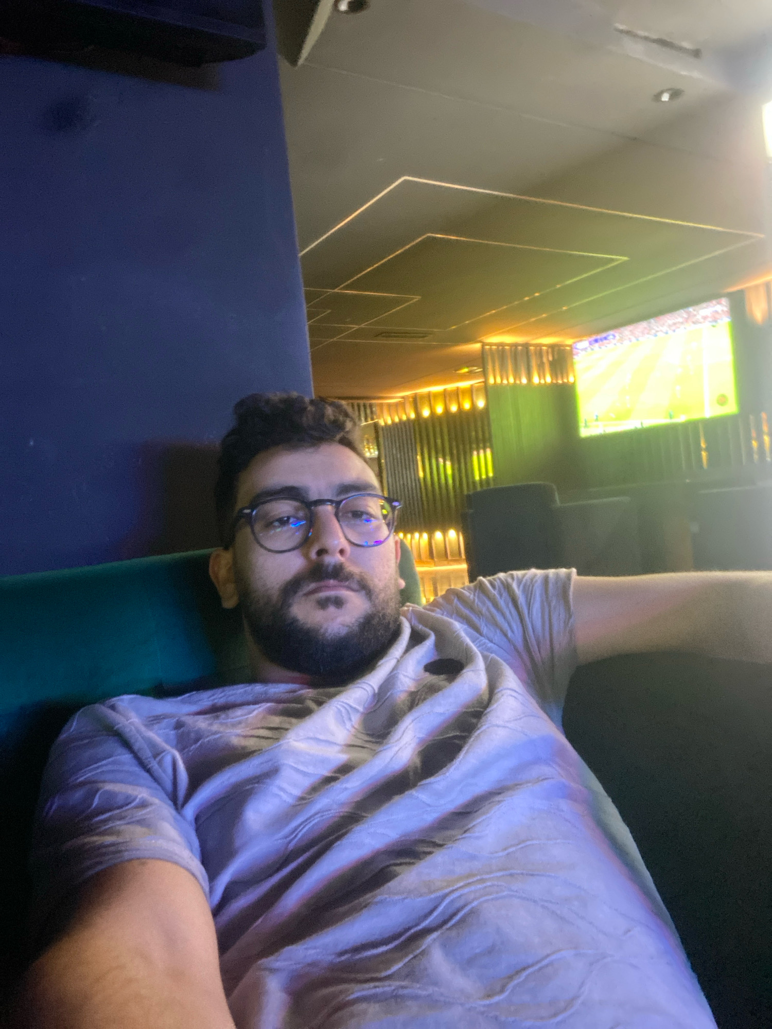 A man with dark hair and a beard, wearing glasses and a light-colored t-shirt with a textured pattern, is taking a selfie. He is seated on a dark green couch in what appears to be a lounge or bar. Behind him, a large screen displays a sports game, likely soccer, with a bright green field and players. The environment features modern lighting with illuminated vertical panels and geometric ceiling designs. The overall ambiance is dimly lit, with the screen and ambient lighting providing the primary illumination. The man is positioned in the foreground, looking directly at the camera. The scene is set in Meknes, Morocco.