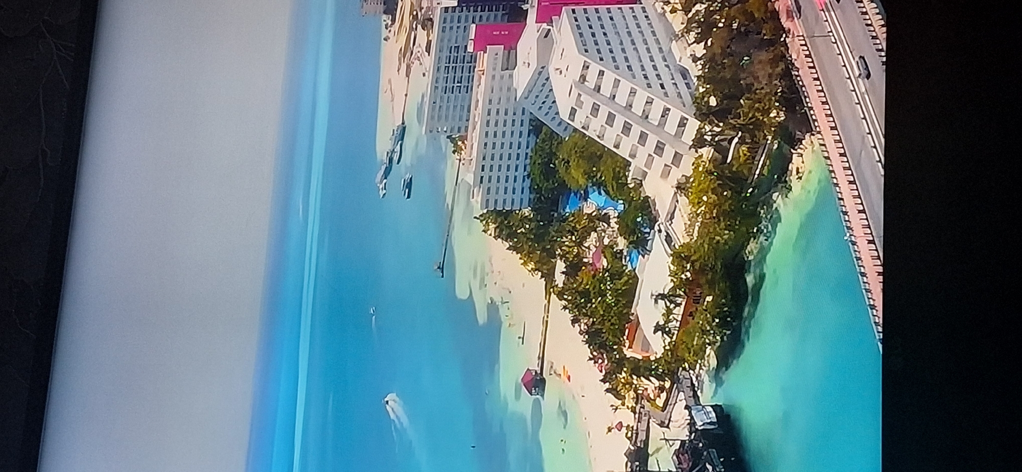 This is an aerial view of a coastal resort area. The ocean is a vibrant blue, with several boats visible on the water, including a speedboat leaving a wake and a ferry or larger vessel docked. Along the shoreline, there is a sandy beach dotted with palm trees and lounge chairs. Several resort buildings are visible, including a large white hotel with many windows, and a unique pyramid-shaped white building. There is also a smaller building with a red roof situated on a pier extending into the water. A swimming pool complex with blue water is nestled among the trees. In the foreground, a road runs alongside the water, with a few cars visible. The sky is a pale blue, merging with the horizon. The image does not depict the city of Bila Tserkva, Ukraine; it appears to be a tropical or subtropical resort location.