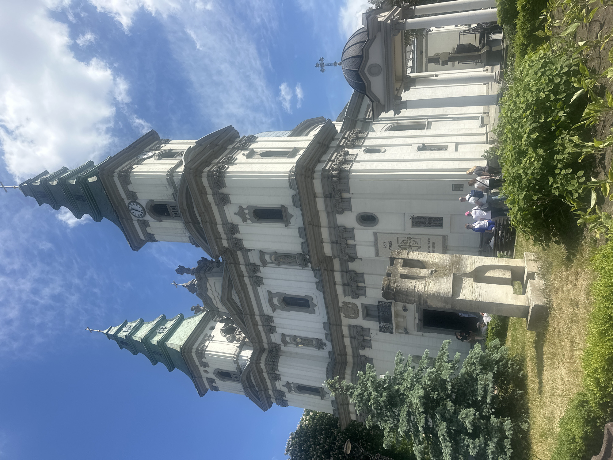 The image shows the Archcathedral Basilica of the Immaculate Conception in Ternopil, Ukraine. The white, baroque-style church features two towers with green roofs, a clock on one tower, and a dome. A stone monument stands in front of the church. People are visible near the entrance. Greenery surrounds the building. The sky is blue with scattered clouds.