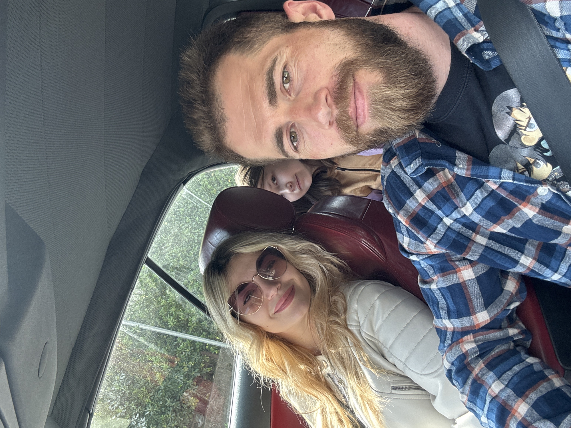 The image is an interior shot of a vehicle, likely a car, with three individuals visible. A man with a beard and wearing a blue plaid shirt over a black t-shirt is positioned closest to the camera. Behind him, partially obscured, is a young girl. In the front passenger seat, a woman with blonde hair and sunglasses is smiling. Red leather seats are visible. The view through the car window suggests an outdoor setting with greenery. The image location is not identifiable from this visual alone. The image is rotated counter-clockwise.