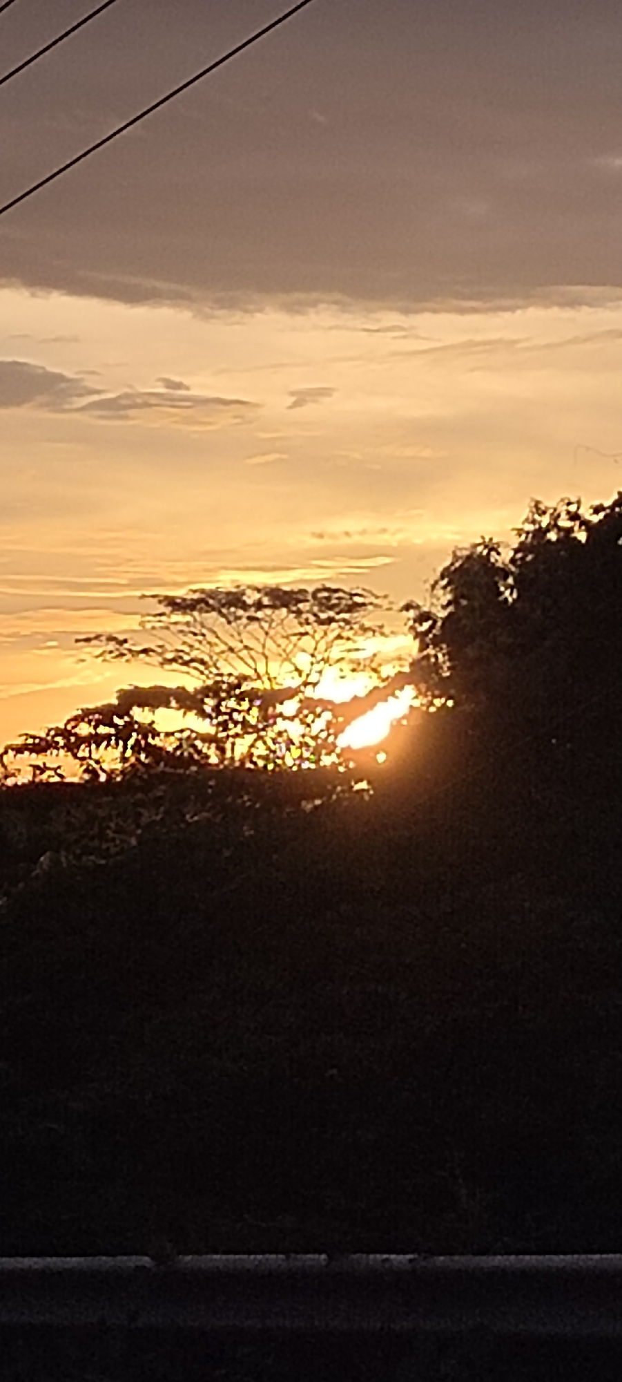 The image captures a sunset in Lhokseumawe, Indonesia. The sun is partially obscured by trees, its rays shining through the branches, creating a bright, warm glow. The sky is a mix of orange, yellow, and grey hues, indicating the time of day. Utility lines are visible in the upper left corner of the frame. The foreground is dark, likely due to dense foliage or shadow. The image appears to have been taken from a low vantage point, possibly near a road or ground level.