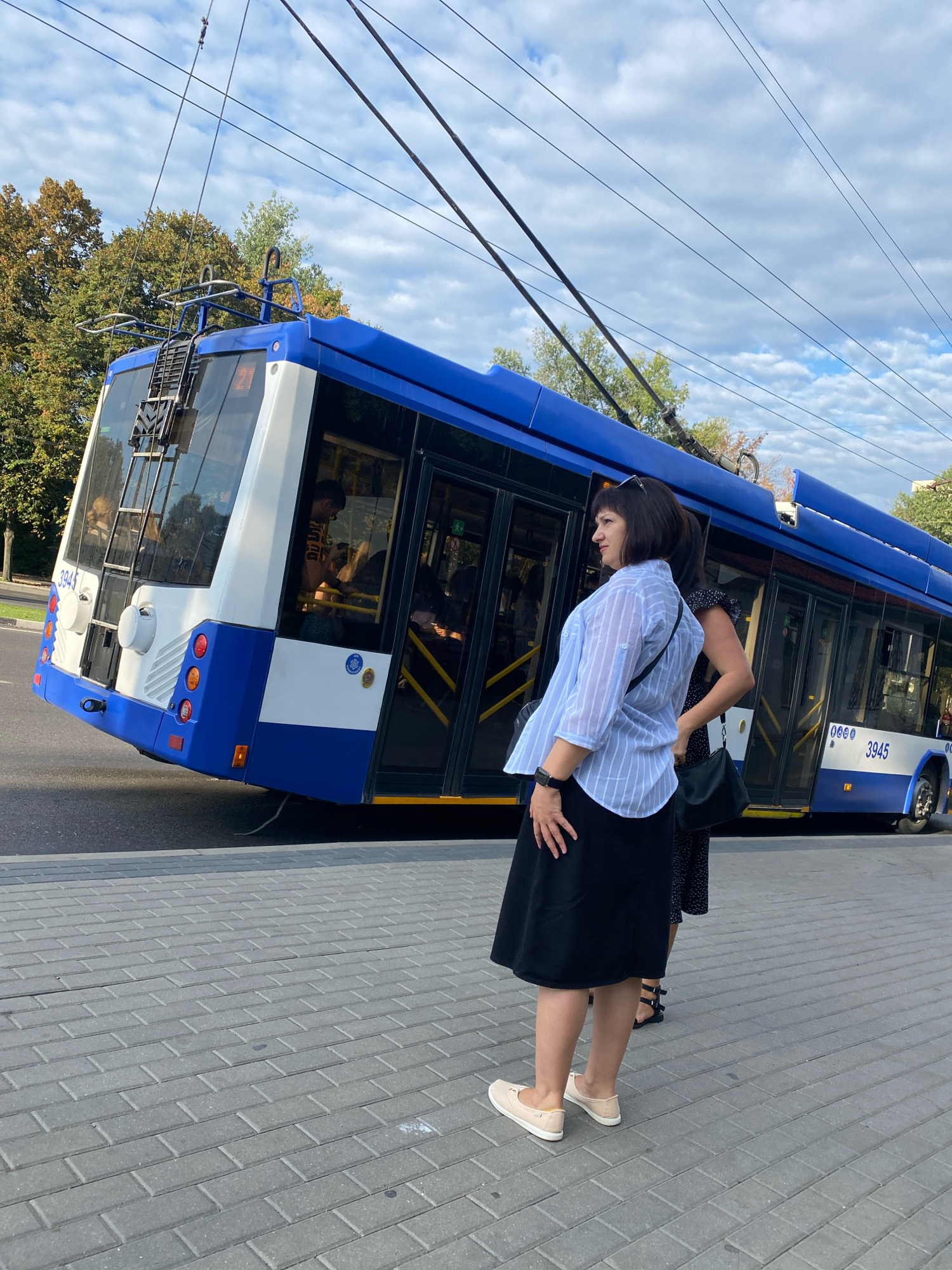 A blue and white trolleybus, number 3945, is partially angled on a street in Chișinău, Moldova, with its front end lifted higher than its rear. The trolleybus is connected to overhead electric wires. In the foreground, a woman with short, dark hair wears a light blue and white striped shirt and a black skirt, standing on a gray brick sidewalk. She is looking to her left. Behind her, another woman in a dark patterned dress is partially visible. Trees with green and yellow foliage line the street behind the trolleybus. The sky is cloudy with patches of blue.