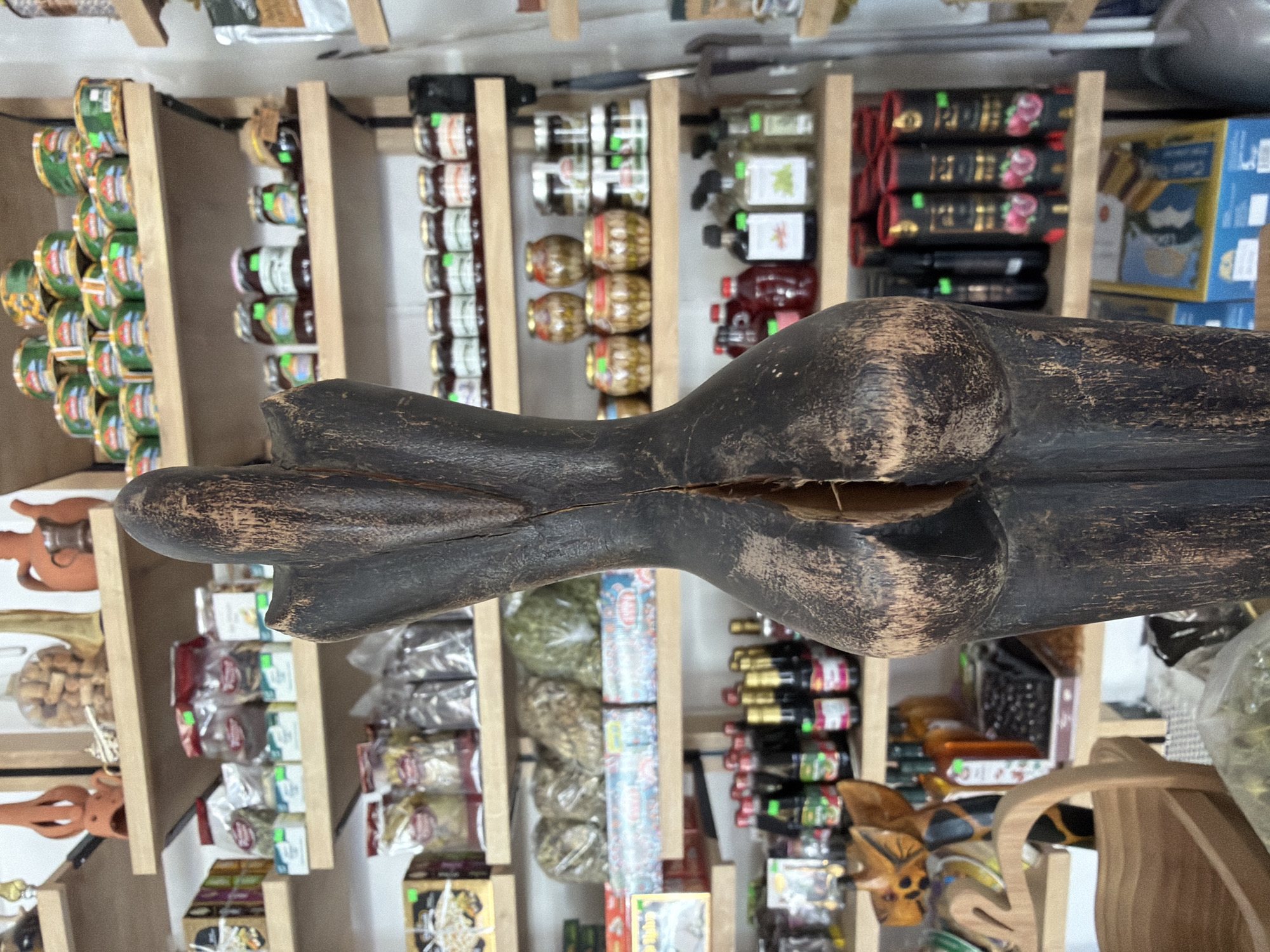 The image is a close-up shot of a dark, wooden, abstract sculpture, possibly representing a female torso, which is positioned in the foreground and takes up a significant portion of the frame. The sculpture has a rough, textured surface with visible wood grain and cracks, notably a prominent vertical split down the center of its posterior.
Behind the sculpture, shelves filled with various products are visible, suggesting a retail environment. These shelves hold an assortment of items, including jars of preserves or spices with colorful labels, bags of dried goods, and bottled liquids such as sauces or oils. Some items are stacked in rows, while others are displayed individually. The shelves themselves are light-colored wood, and the overall background suggests a shop interior.
Specific items on the shelves include rows of small jars with red lids on the second shelf from the top. The third shelf down displays several round, golden-colored jars, some with red lids, and a few taller, clear jars with green labels. Further down, there are rows of dark bottles with red and white labels, as well as bottles with green labels. On the bottom shelves, there are cylindrical containers with images of berries and boxed items.
The lighting appears to be natural or from overhead store lights, illuminating the products on the shelves and casting a shadow behind the sculpture. The depth of field is shallow, with the sculpture in sharp focus and the background items progressively blurring.
Based on the context provided by the user, this scene is located in a shop in Marmaris, Turkey. The merchandise visible on the shelves, such as preserved goods and possibly local crafts or food products, is consistent with what one might find in a Turkish market or specialty store. The wooden sculpture itself could be a decorative item or a representation of local art.