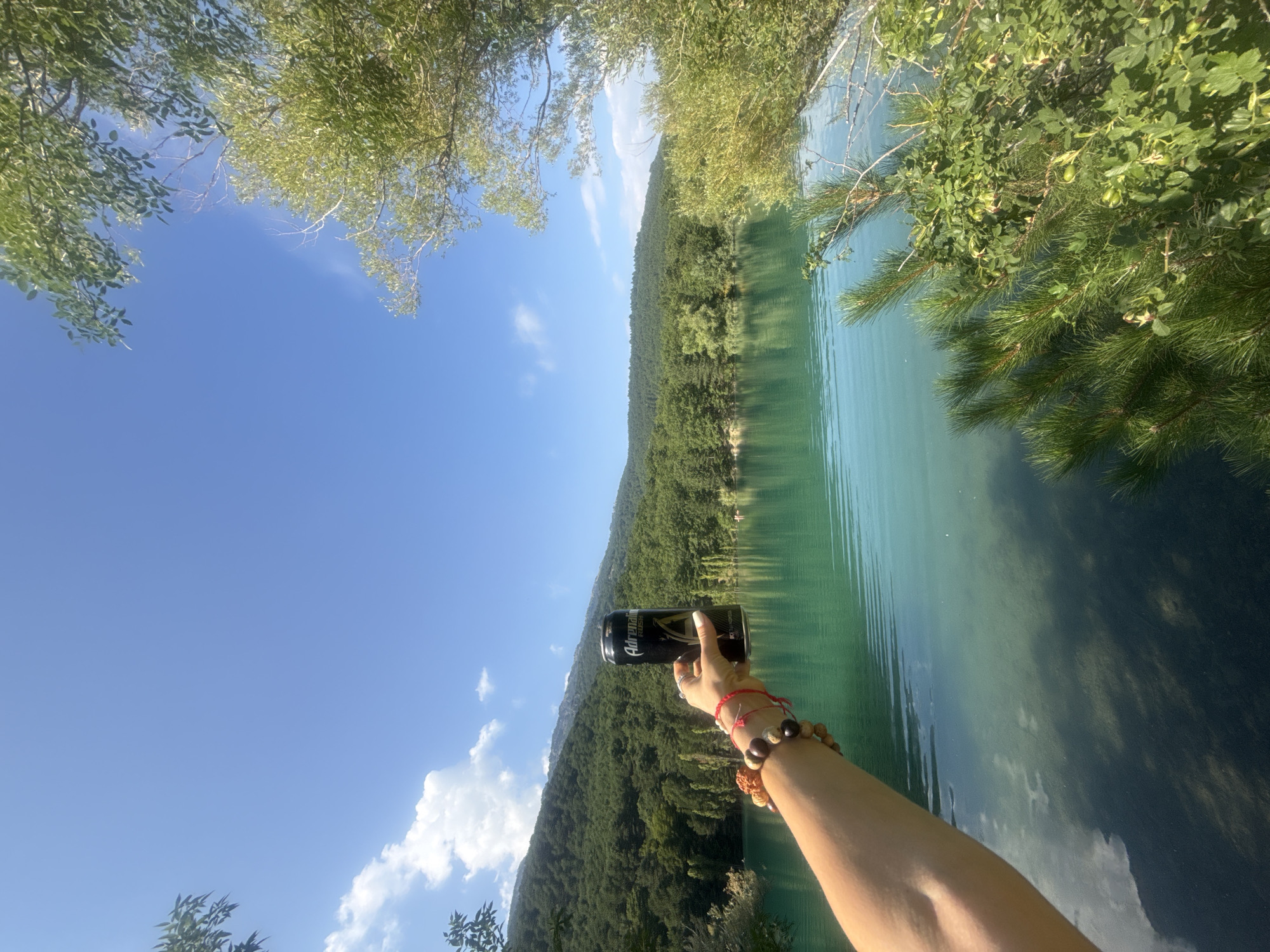 A person's arm, adorned with several bracelets, extends into the frame from the left, holding a black can of "Adrenaline Rush" energy drink. The can has a yellow emblem and Russian text. The scene is set by a large, clear turquoise lake with gentle ripples. In the background, a dense forest covers rolling hills that rise to meet a bright blue sky with scattered white clouds. The lighting suggests it is daytime, likely mid-morning or afternoon, with the sun casting a gentle glow on the water and landscape. The location is identified as Unknown, Ukraine.