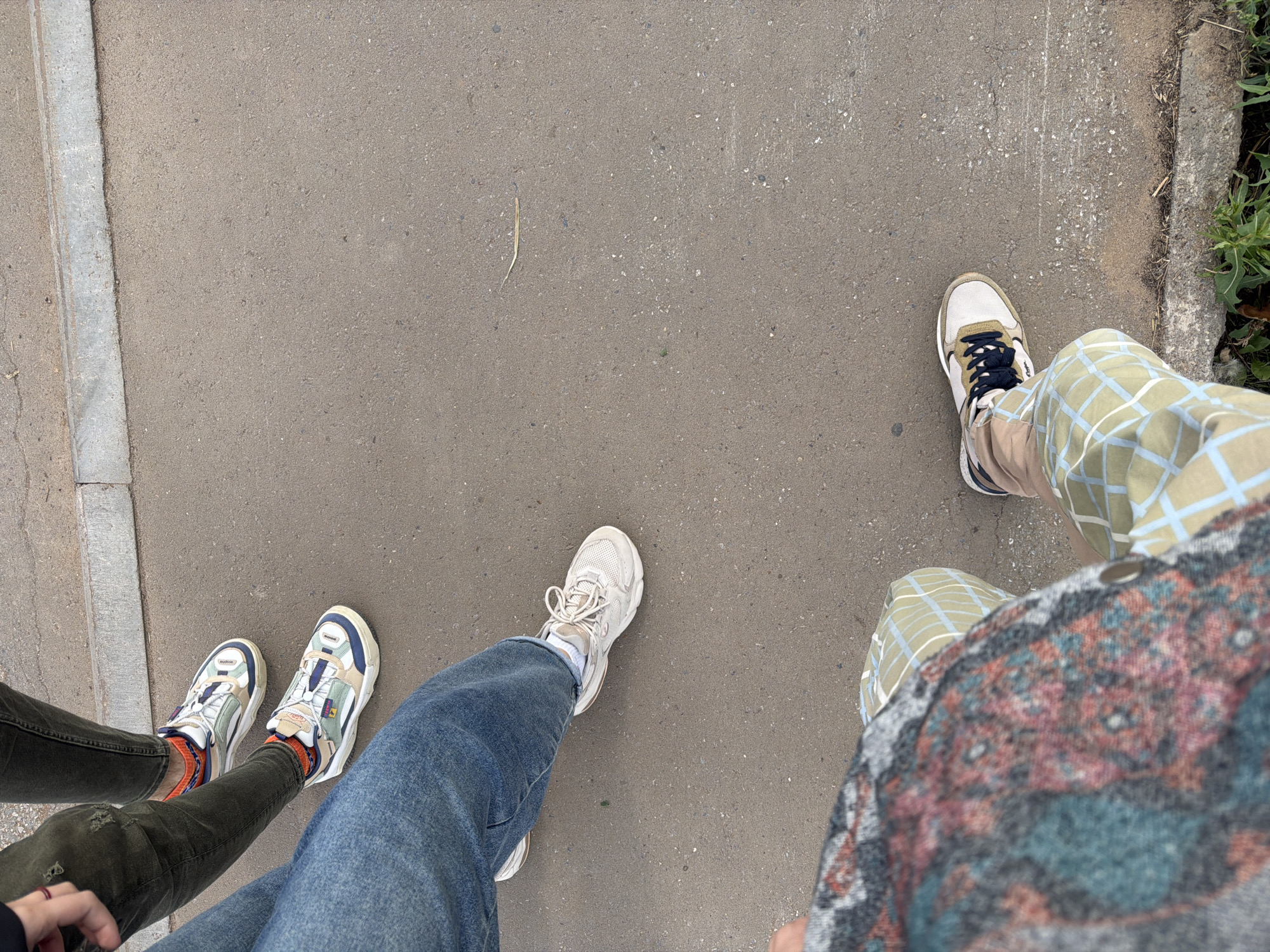 The image shows a ground-level view of people's legs and feet as they stand or walk on an asphalt surface near a concrete curb. The asphalt is mottled with small stones and appears worn. In the background, a concrete curb separates the asphalt from a lighter-colored, possibly paved, surface. A single dry blade of grass lies on the asphalt.
Three pairs of feet are visible, each wearing different footwear and attire. The leftmost pair of feet are clad in dark green trousers and white sneakers with colorful accents, including orange socks with a patterned band. The middle pair of feet are in blue denim jeans and light-colored sneakers. The rightmost pair of feet are in light brown pants with a faint plaid pattern and sneakers with white, tan, and dark blue elements. The clothing suggests a casual outdoor setting. There is no visual information in the image that allows for an identification of the specific location within Yelabuga, Russia.